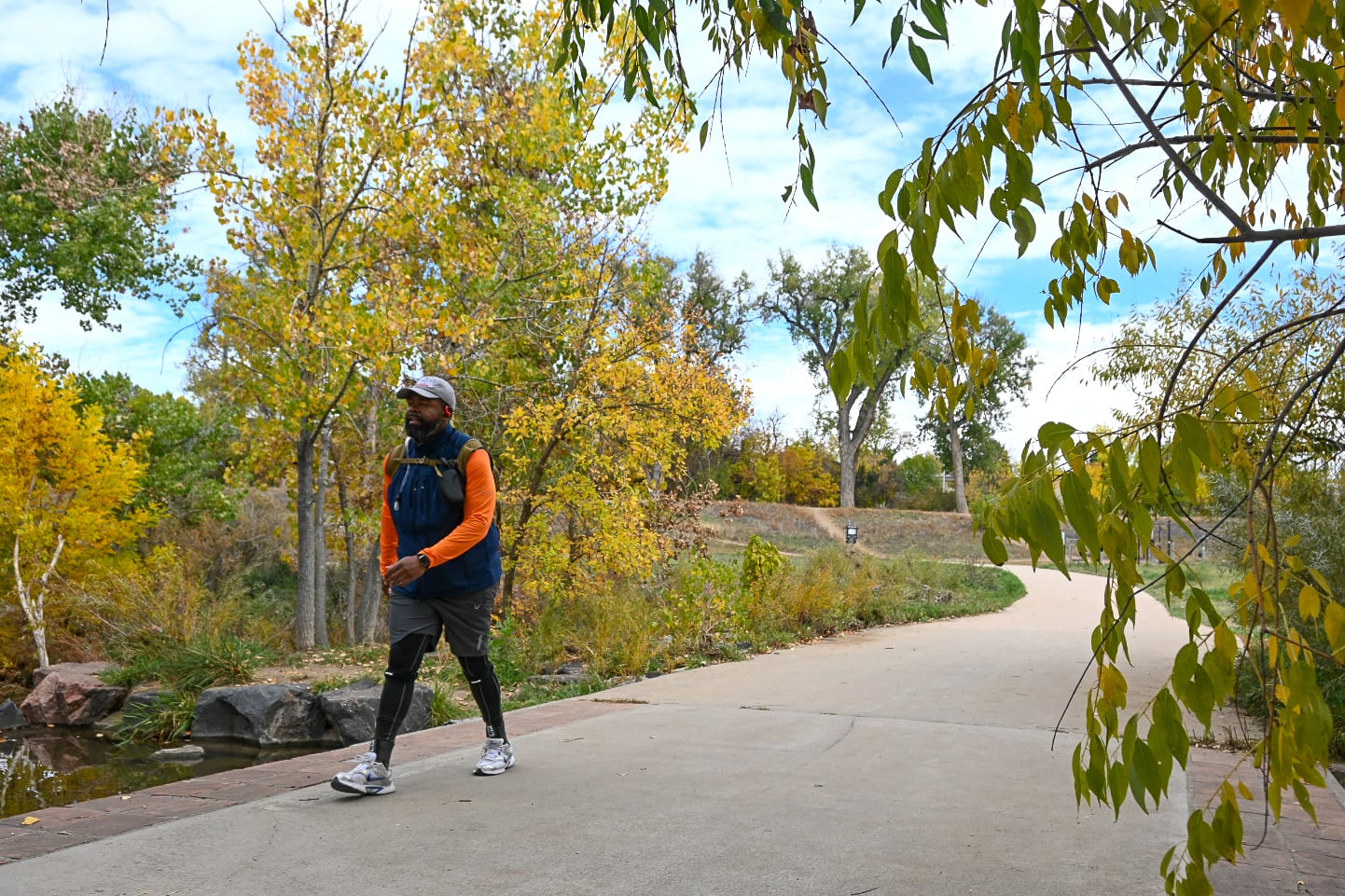 A man in a baseball cap, orange shirt and dark vest walks on a path.