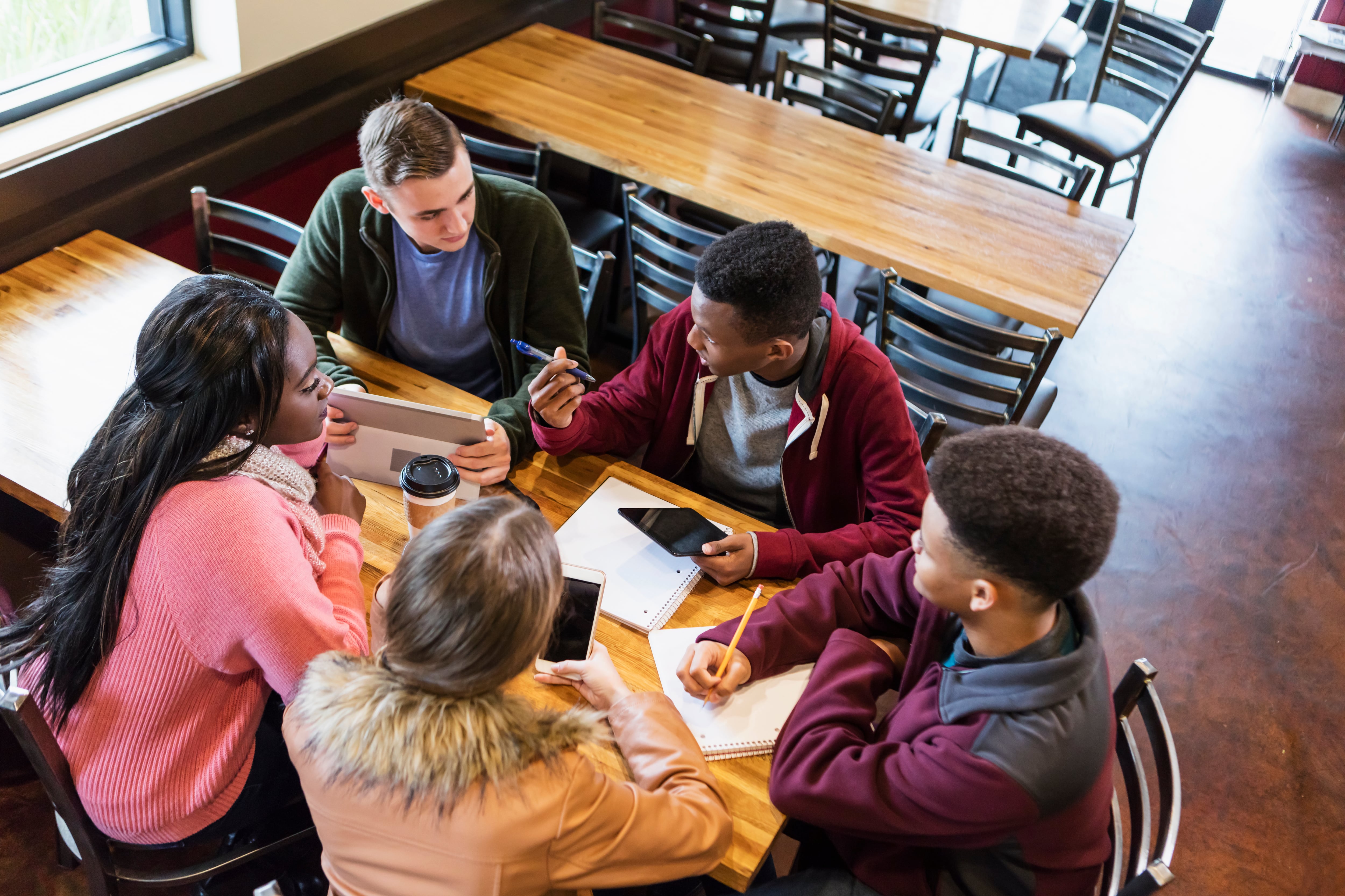 Five teenagers are seated around a table in a coffee shop working together with notebooks, cell phones, and a laptop in view.
