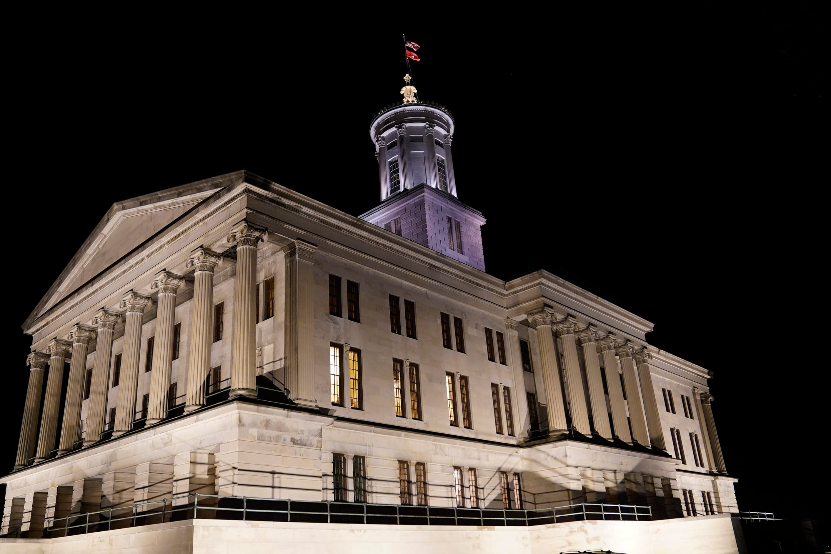 An illuminated large, tan stone building with a flag at the top with a dark night sky in the background.