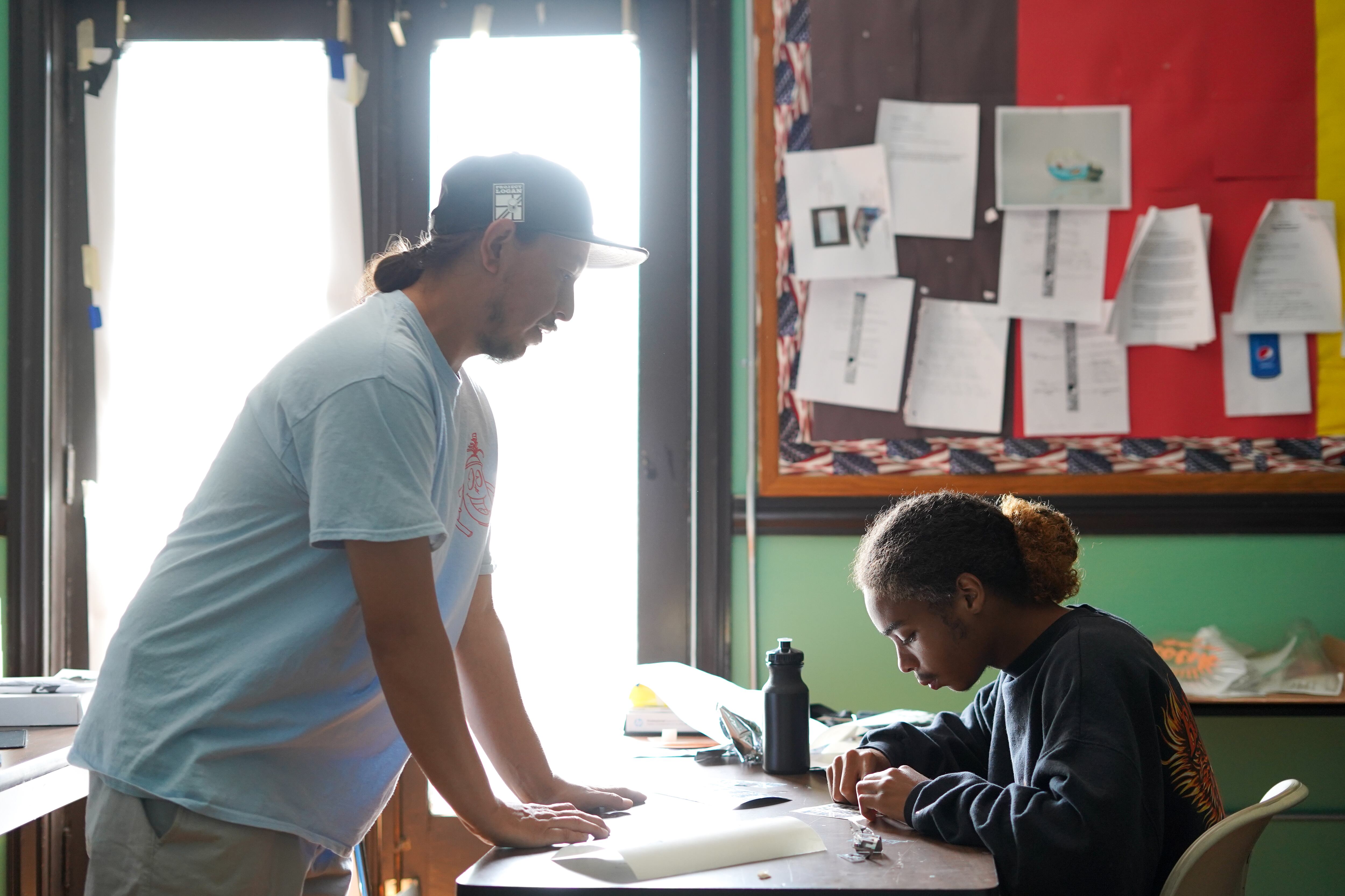 A teacher stands in front of a student while instructing the student on a task. The student is sitting down at a desk while working. The student and teacher and working in classroom, in front of a large window.