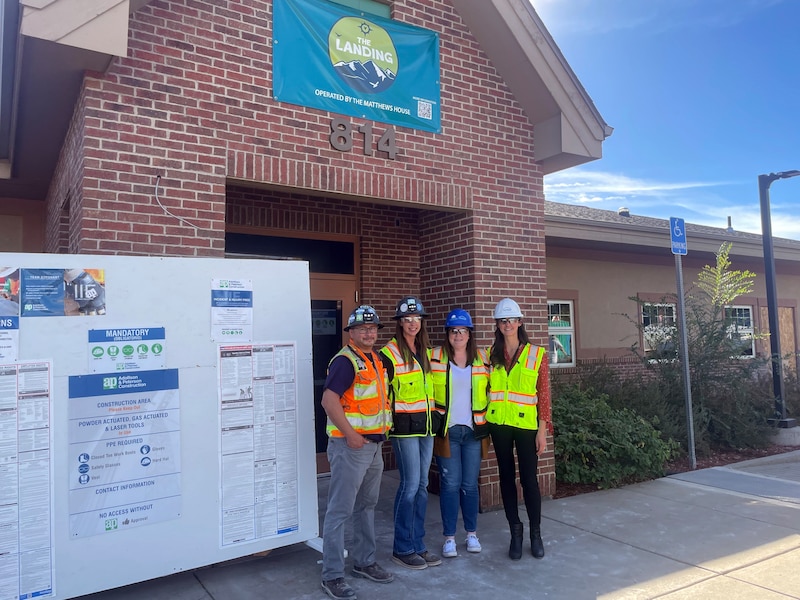 Four people in hard hats and safety vests pose for a photograph outside of a building next to a giant poster.