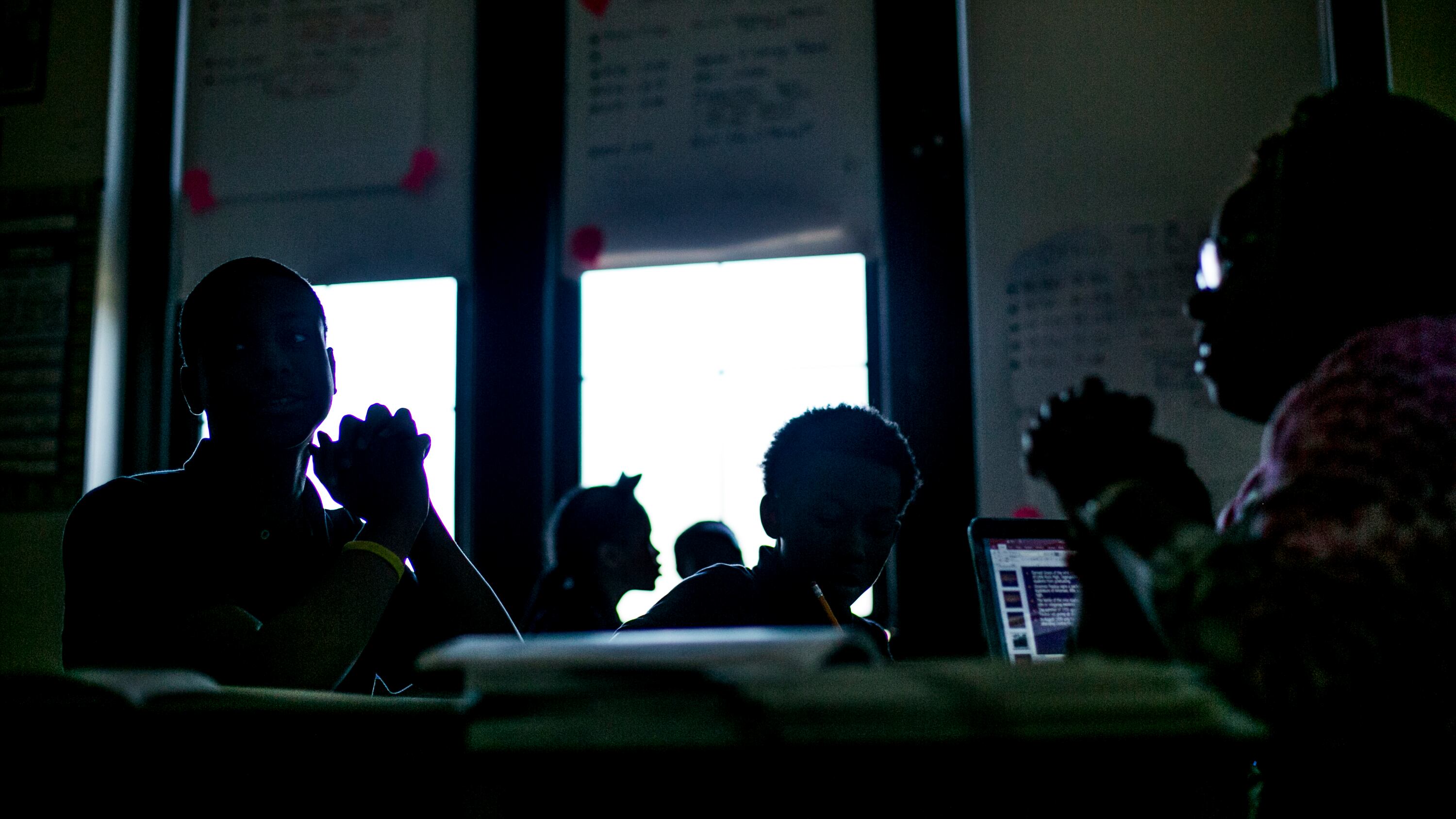 Students and their teacher sit in a dark classroom, the students silhouetted against the windows.