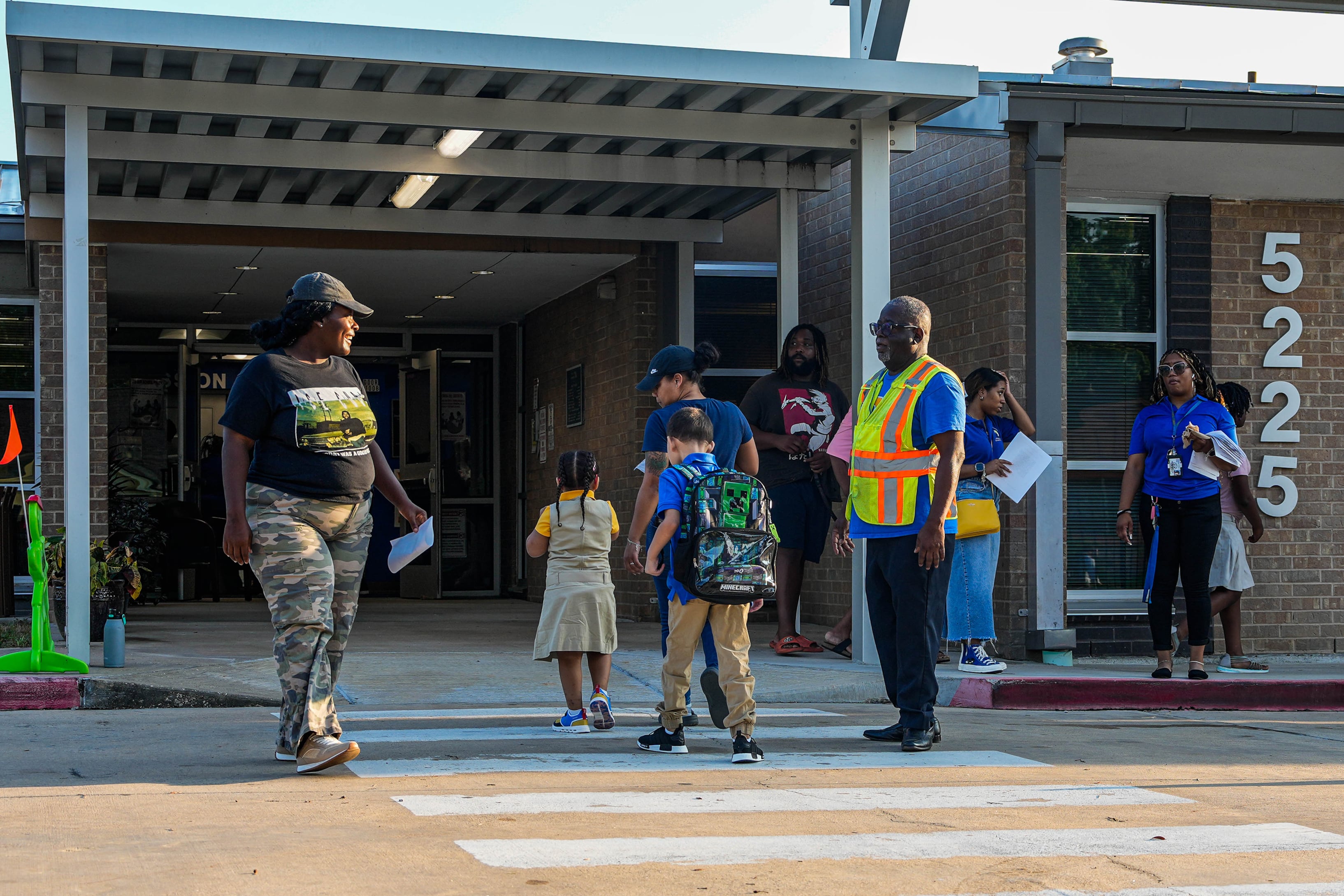 Adults and students cross the street at a cross walk with a school building in the background.