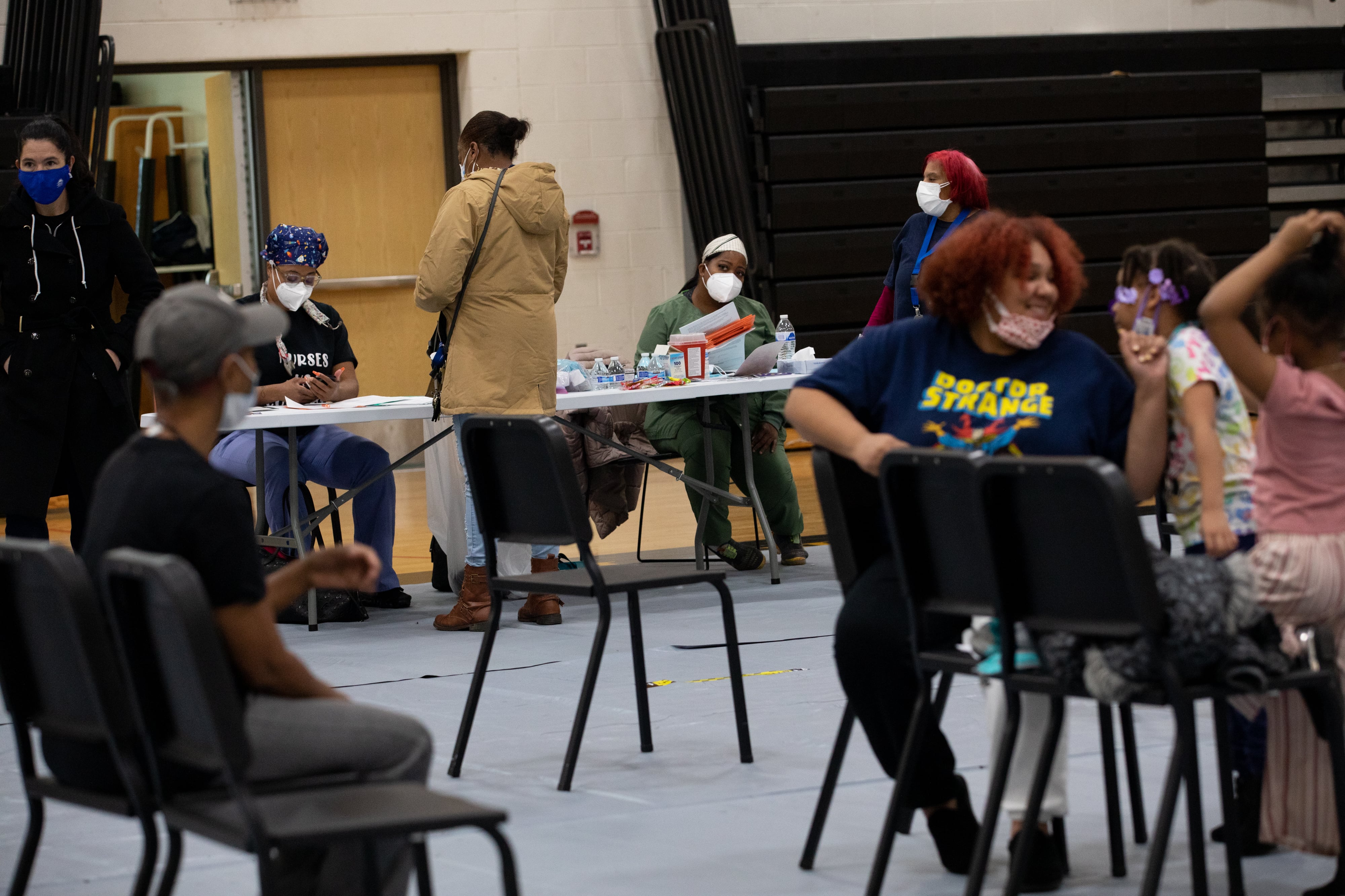 A group of masked parents and children are in a school gymnasium receiving vaccine shots.