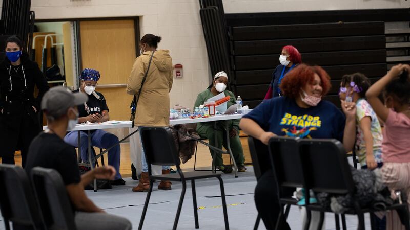 A group of masked parents and children are in a school gymnasium receiving vaccine shots.