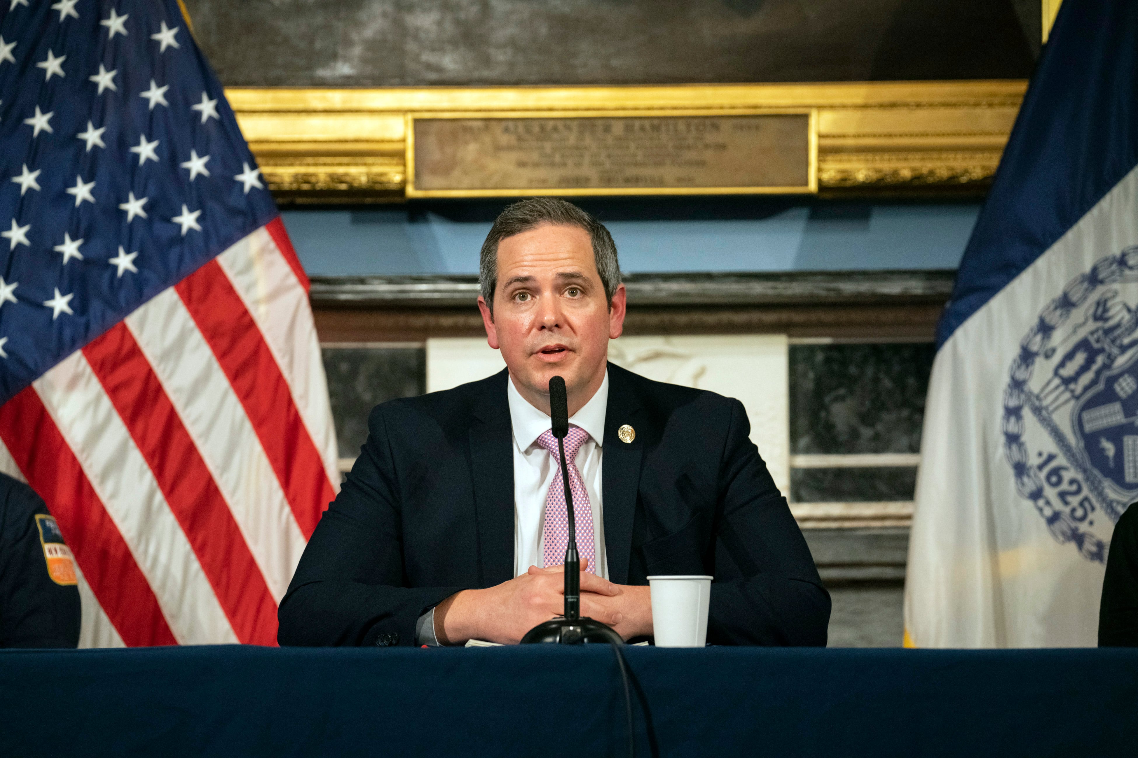 A man in a dark suit and wearing a red tie sits behind a microphone at a table between an American and a NY State flag.