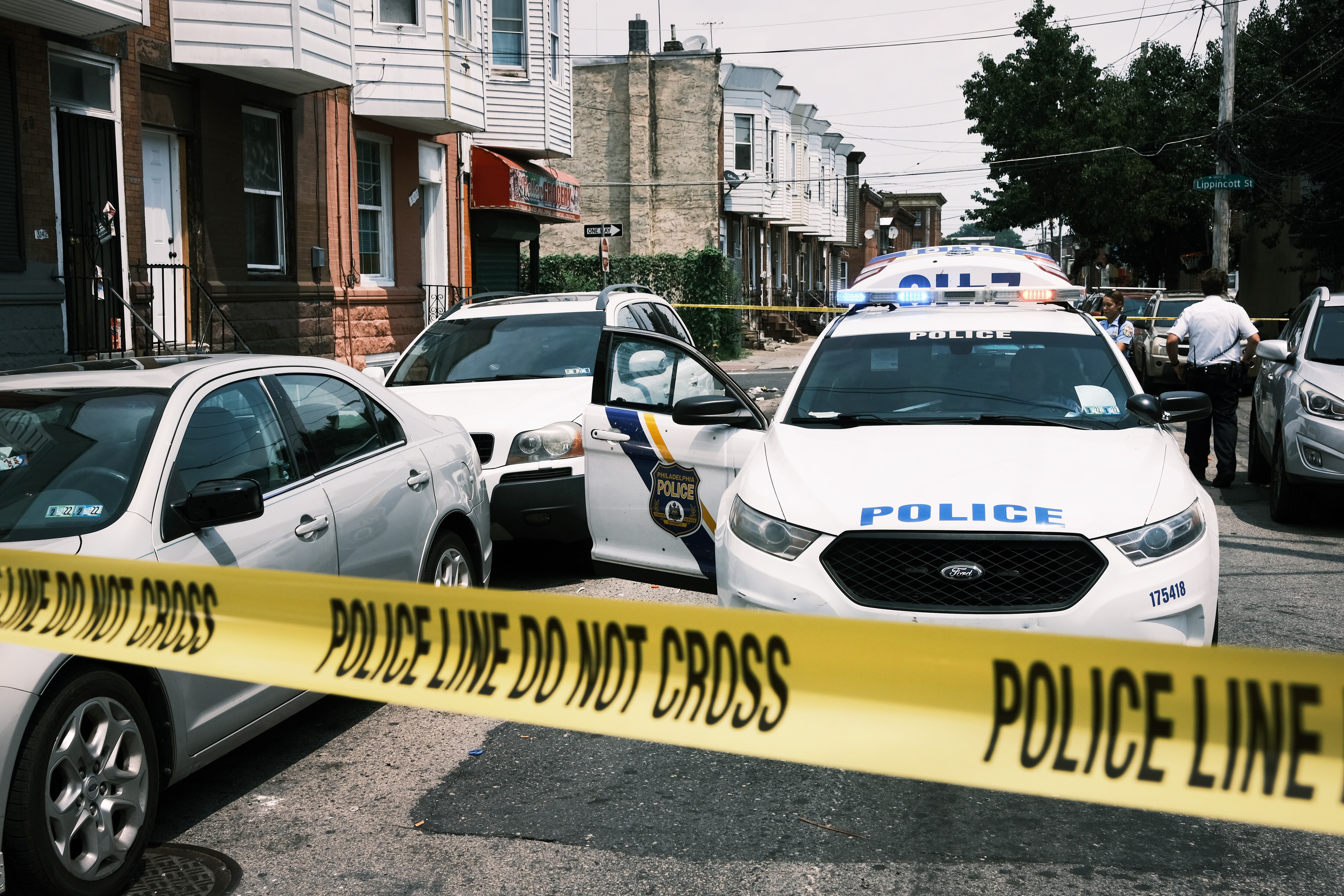 Yellow police tape blocks off a city street. Behind it, are parked cars to the left and a police car in the center.