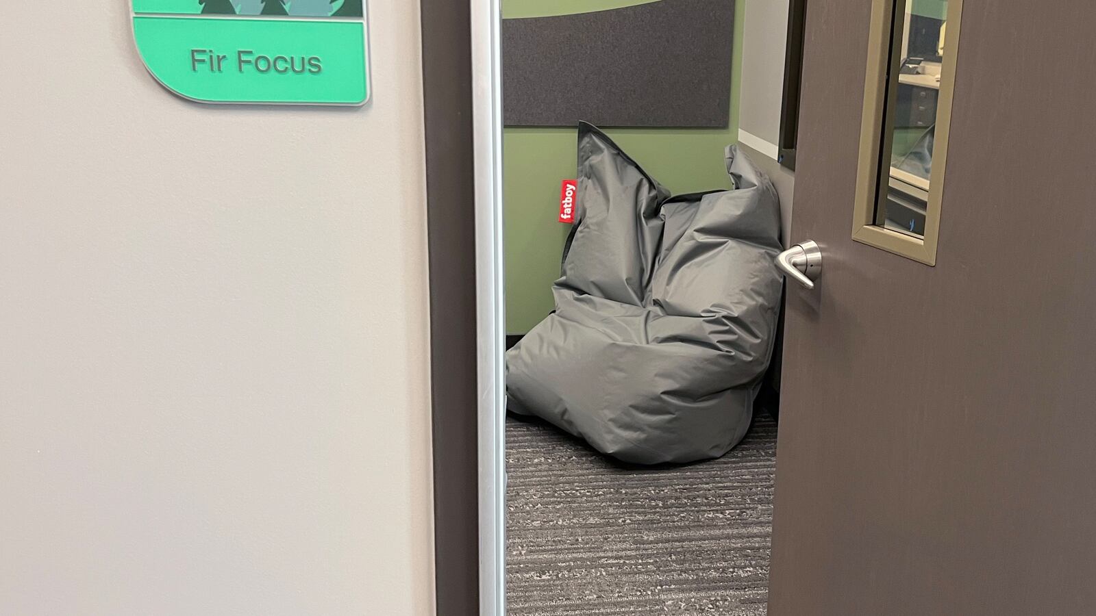A grey wall and brown door open up to a bean bag that sits on the floor in the corner of the room.  
