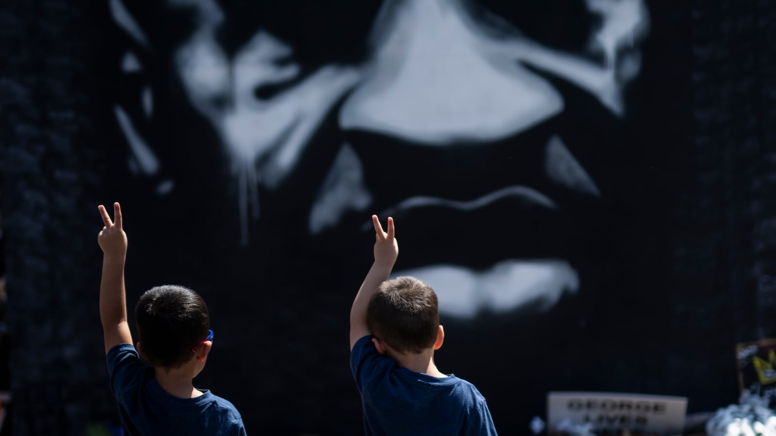 Two little boys, wearing similar blue shirts and shorts, wave a peace sign to a mural of George Floyd. There are rows of flowers between them and the mural.