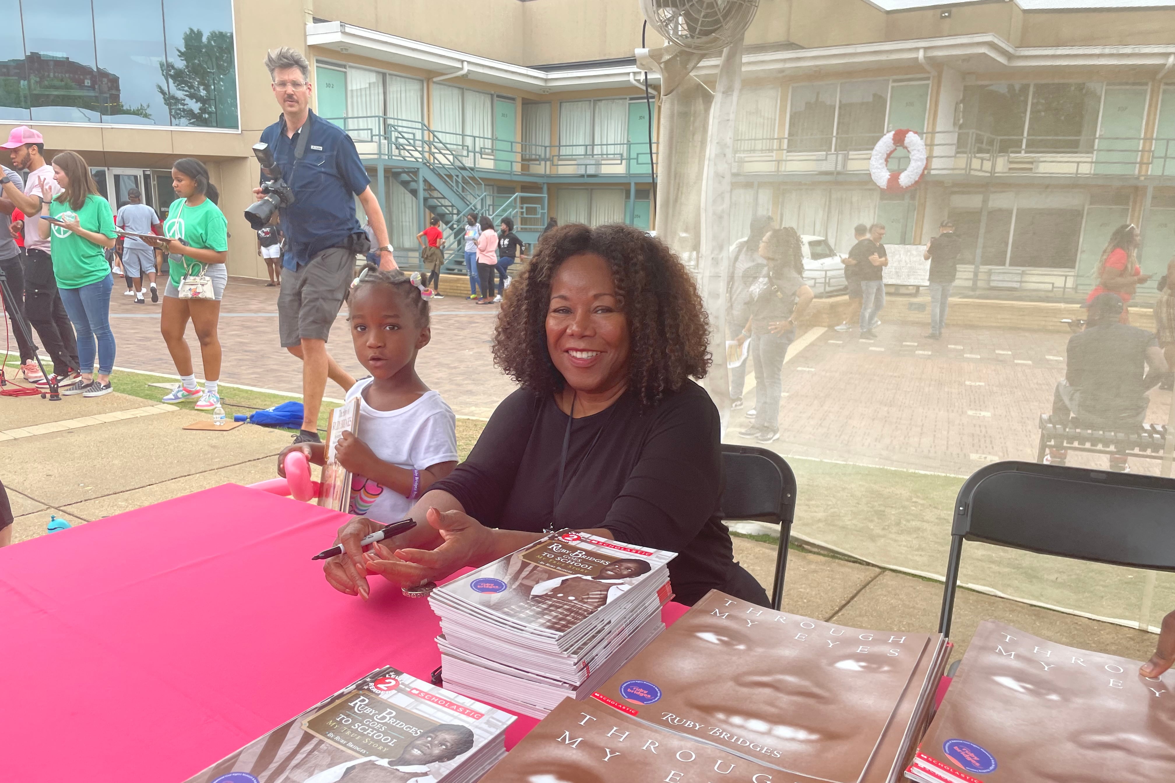 A woman seated at a table with stacks of books featuring an image of a child on the cover. A small child is behind the woman.
