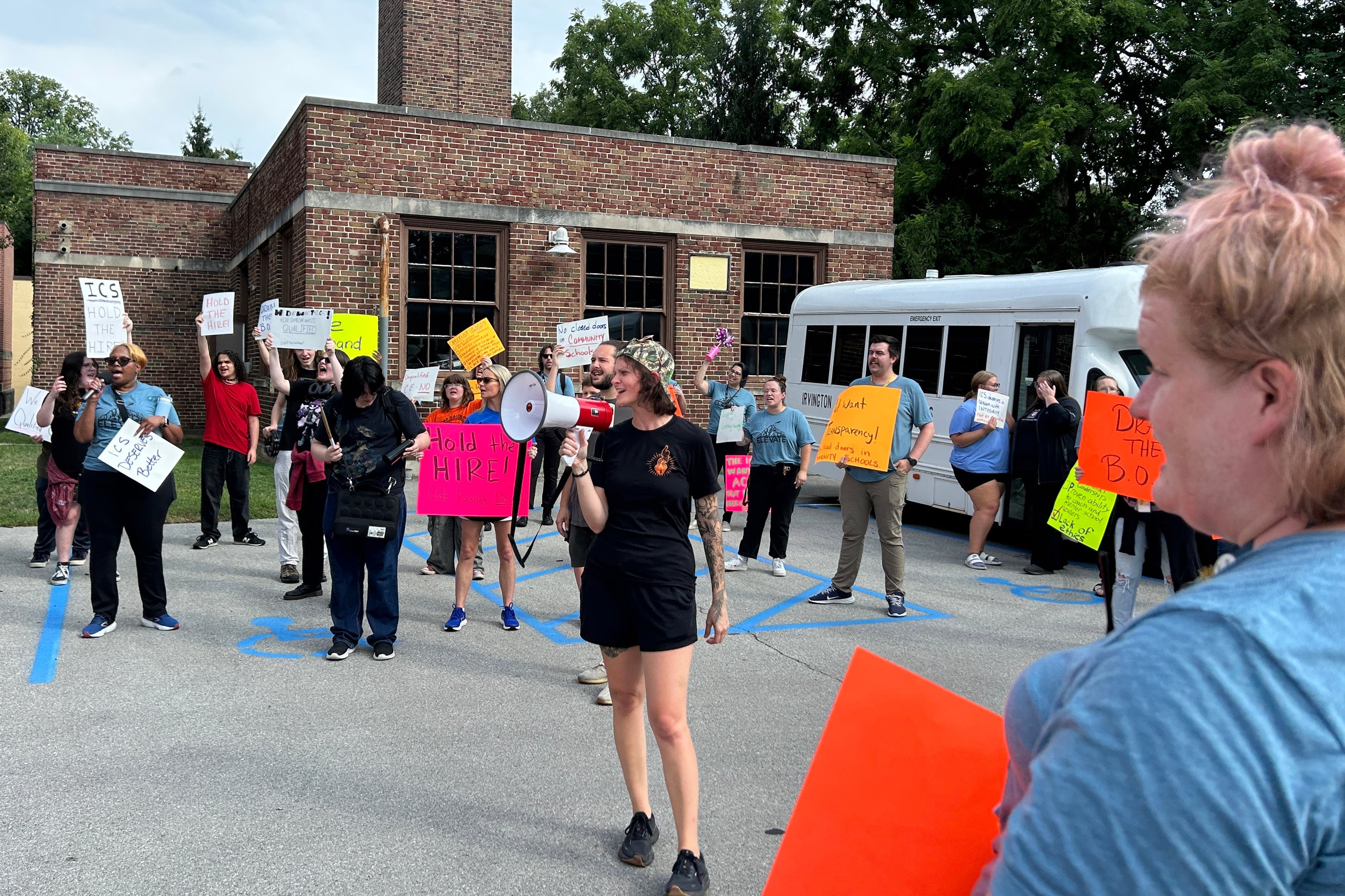A photograph of a large group of protestors, some holding large neon paper signs stand outside.