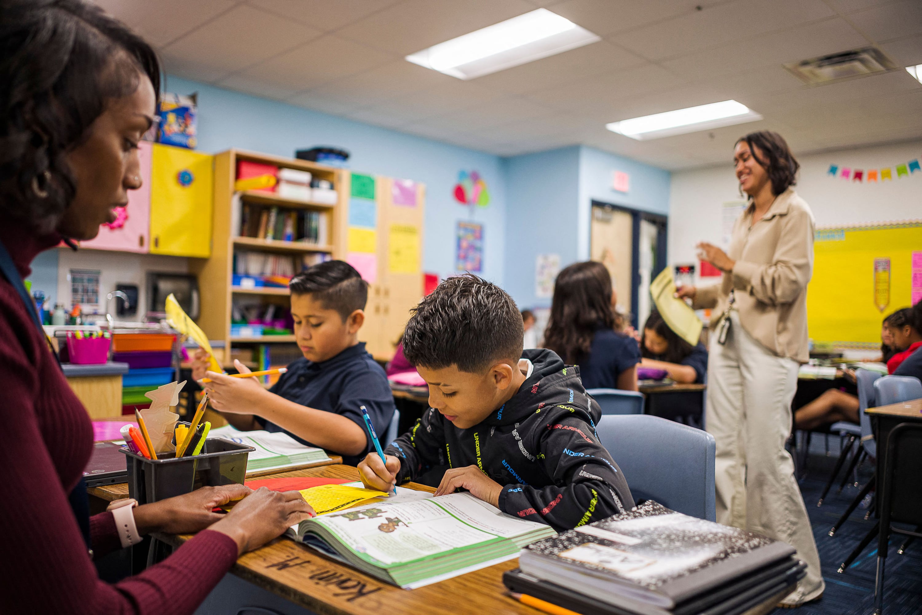 Two adults and three students work in a colorful classroom with blue walls in the background.