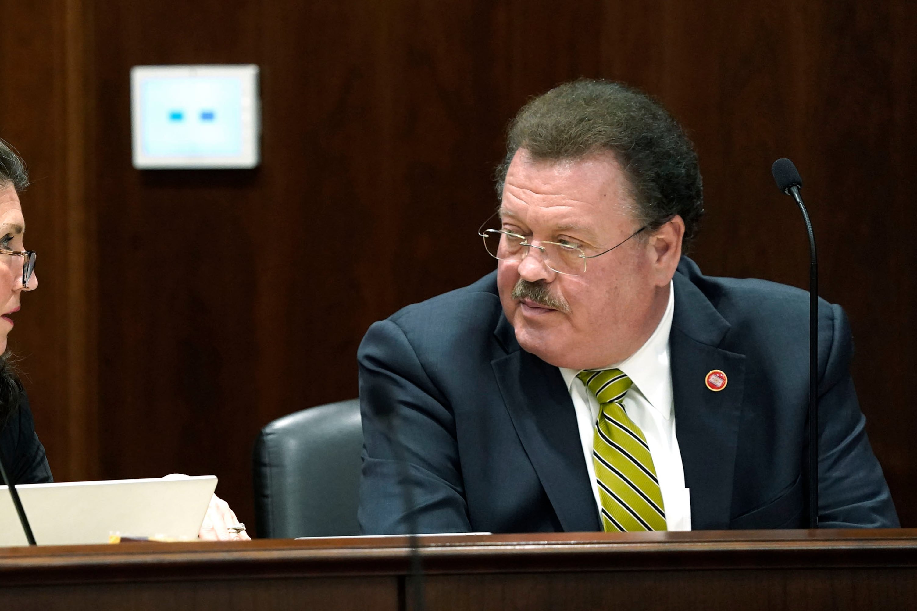 A man wearing a suit speaks with a woman in a paneled committee room