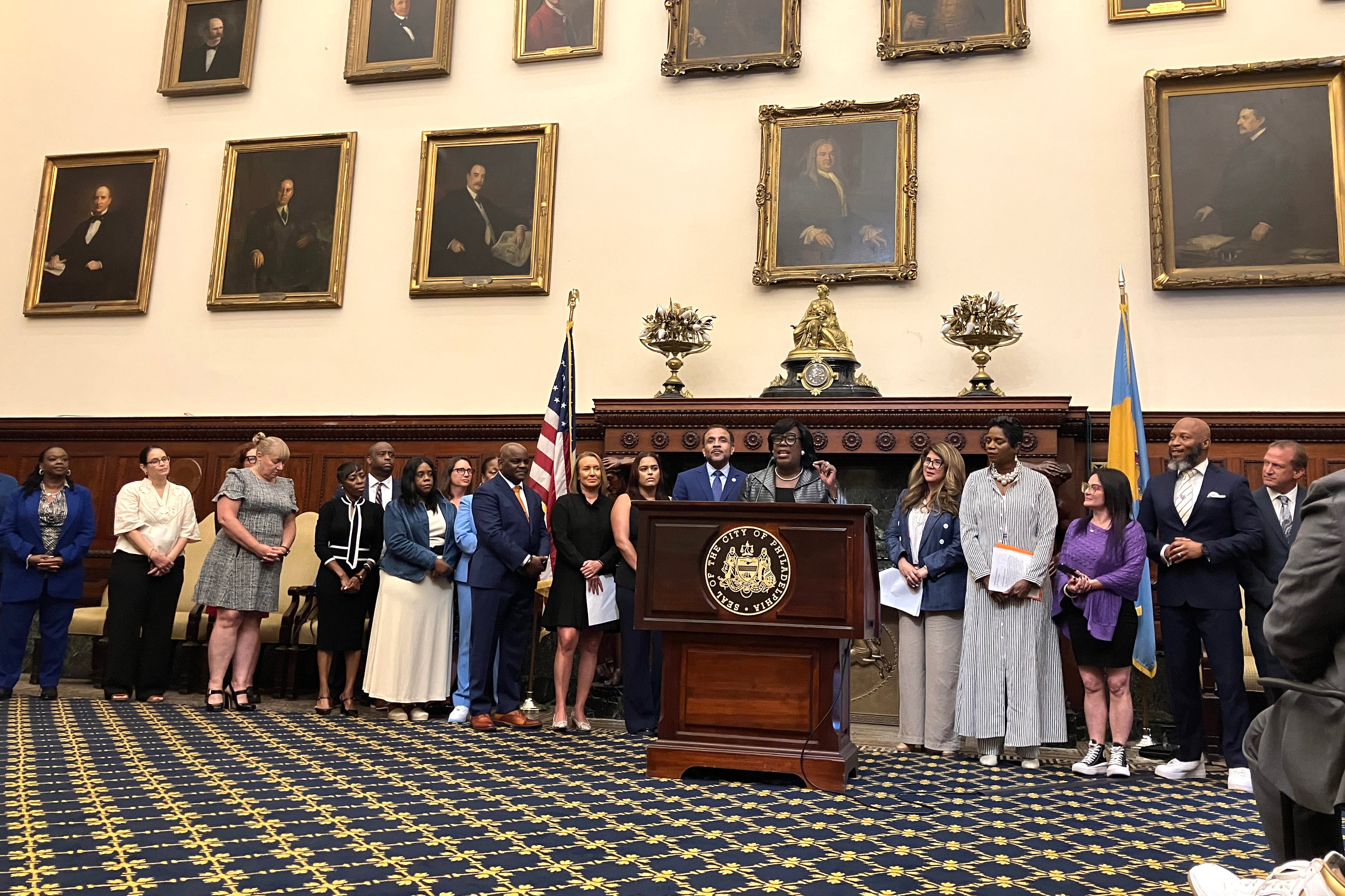 People line up behind a podium and in front of old paintings on the wall.