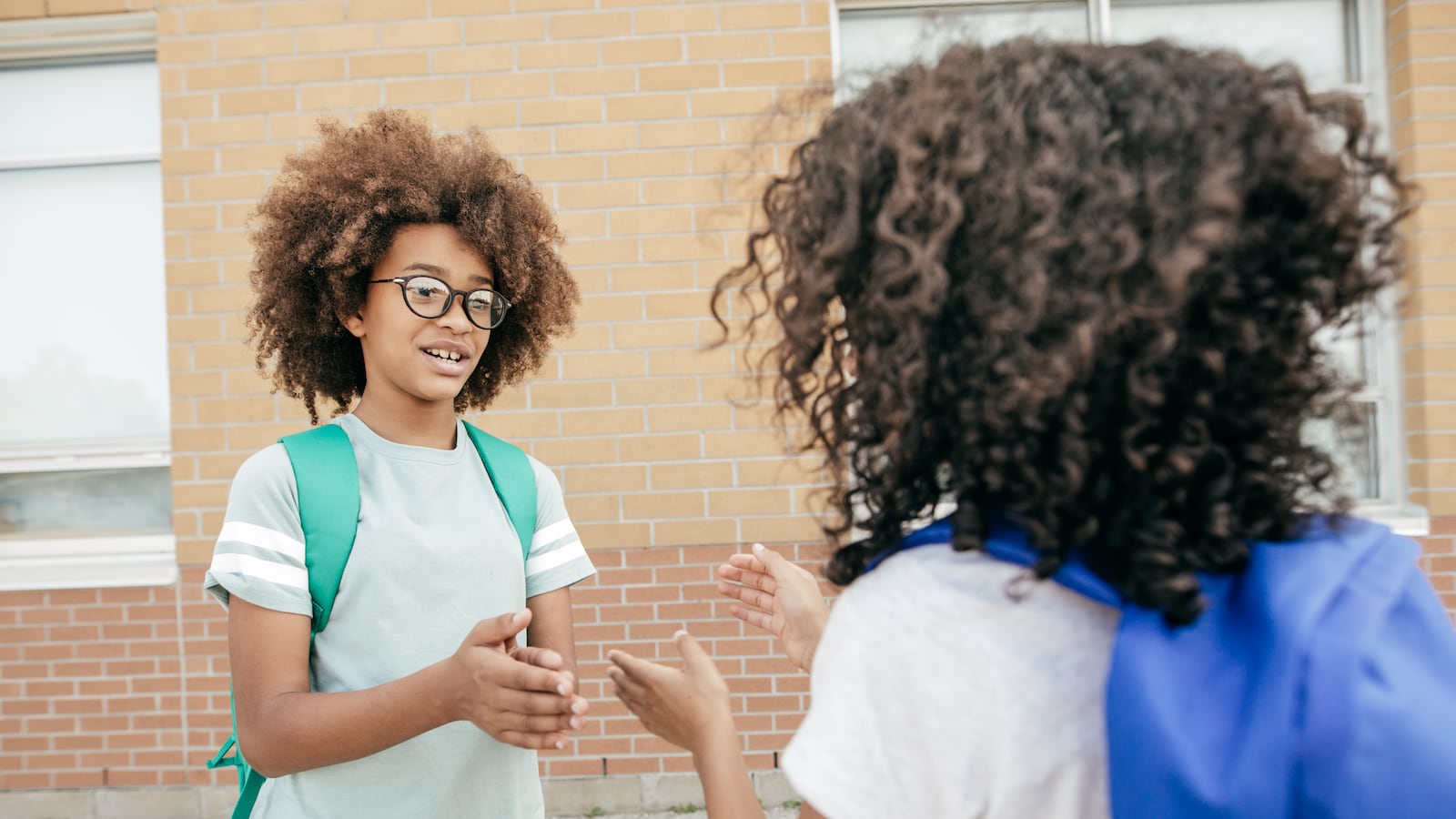 Two students play a game together outside a school building. One is wearing a blue backpack and one is wearing a green backpack.