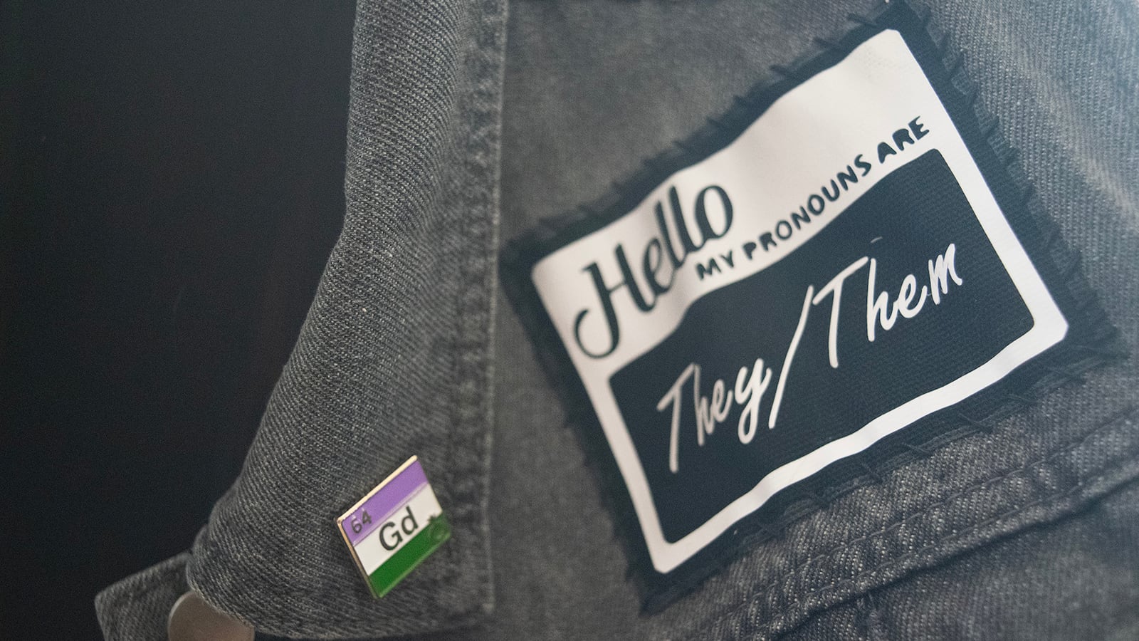 On a gray vest is a patch that reads “Hello my pronouns are they/them.” A small square pin with the genderqueer flag is pinned on the lapel. The pin is in the shape of a periodic table square.