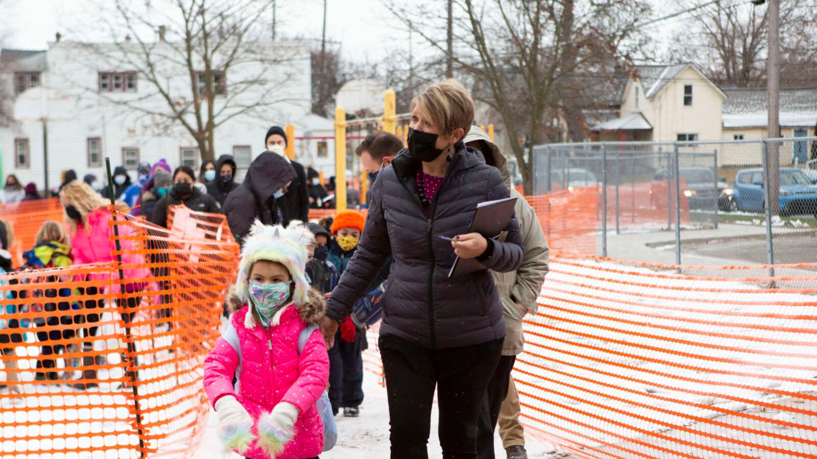 A student in a pink coat and fuzzy hat and mask is led in school by a masked teacher with a line of students following.