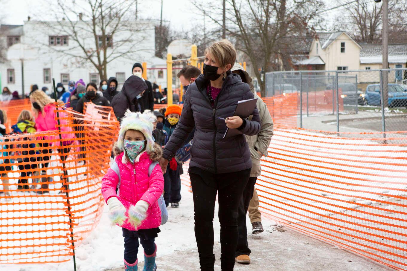 A student in a pink coat and fuzzy hat and mask is led in school by a masked teacher with a line of students following.