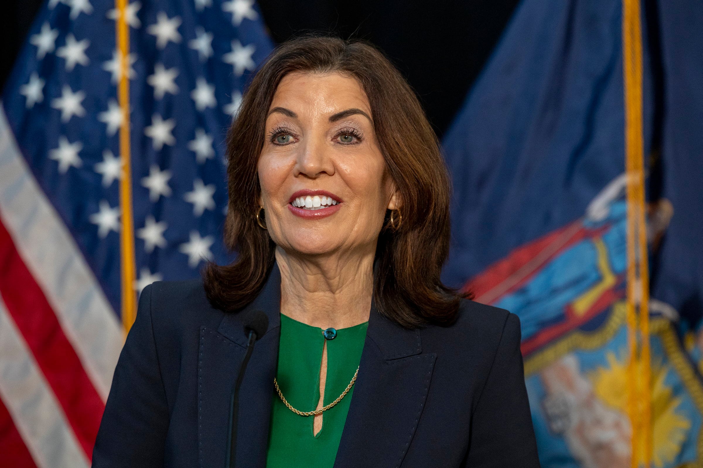 A woman with short dark hair and wearing a dark blue blouse stands in front of two flags.