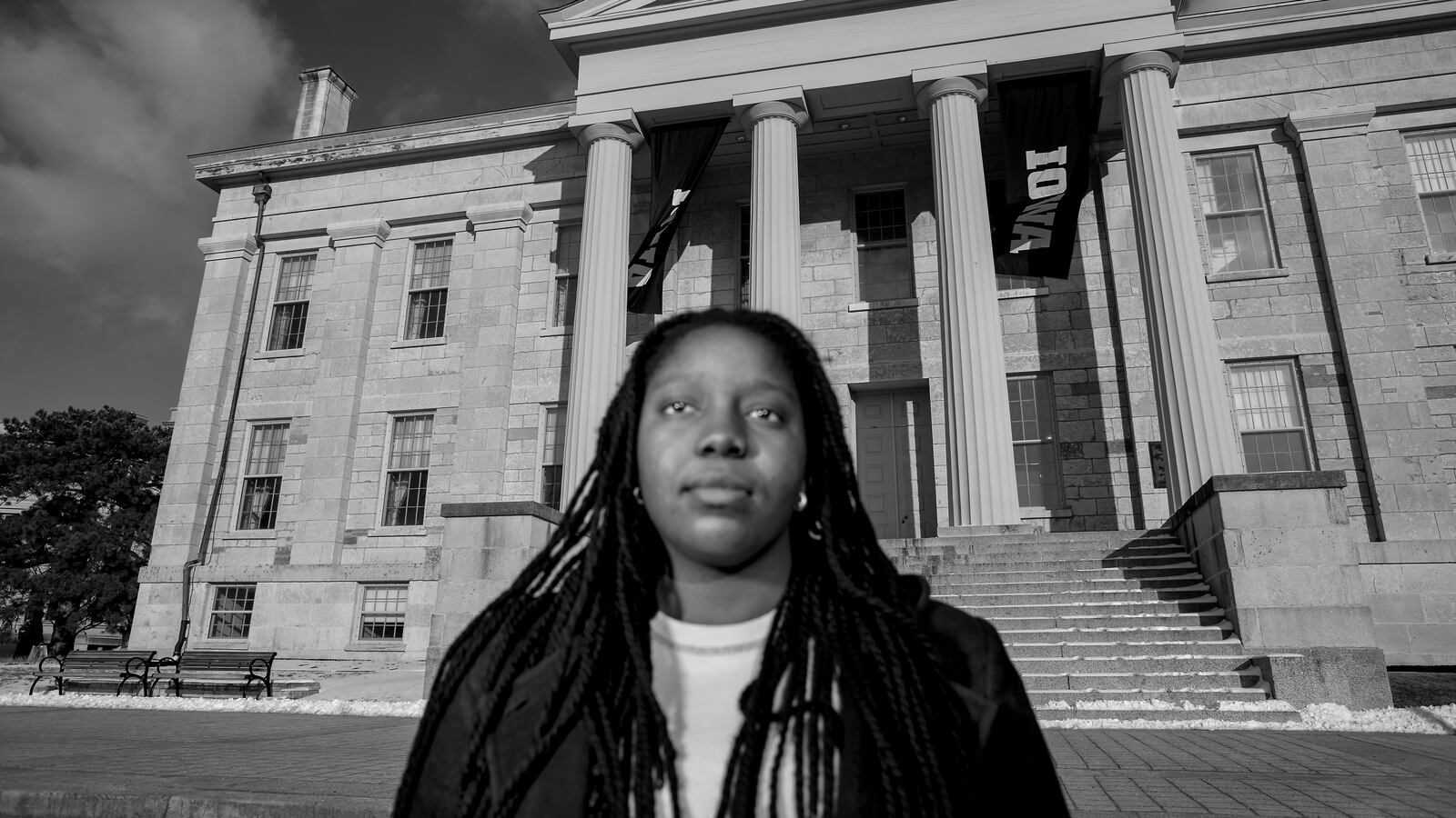 In a black-and-white photograph, young woman with long, black braided hair stands in front of a large building with stone columns. The focus is on the building with the woman slightly out-of-focus in the foreground.