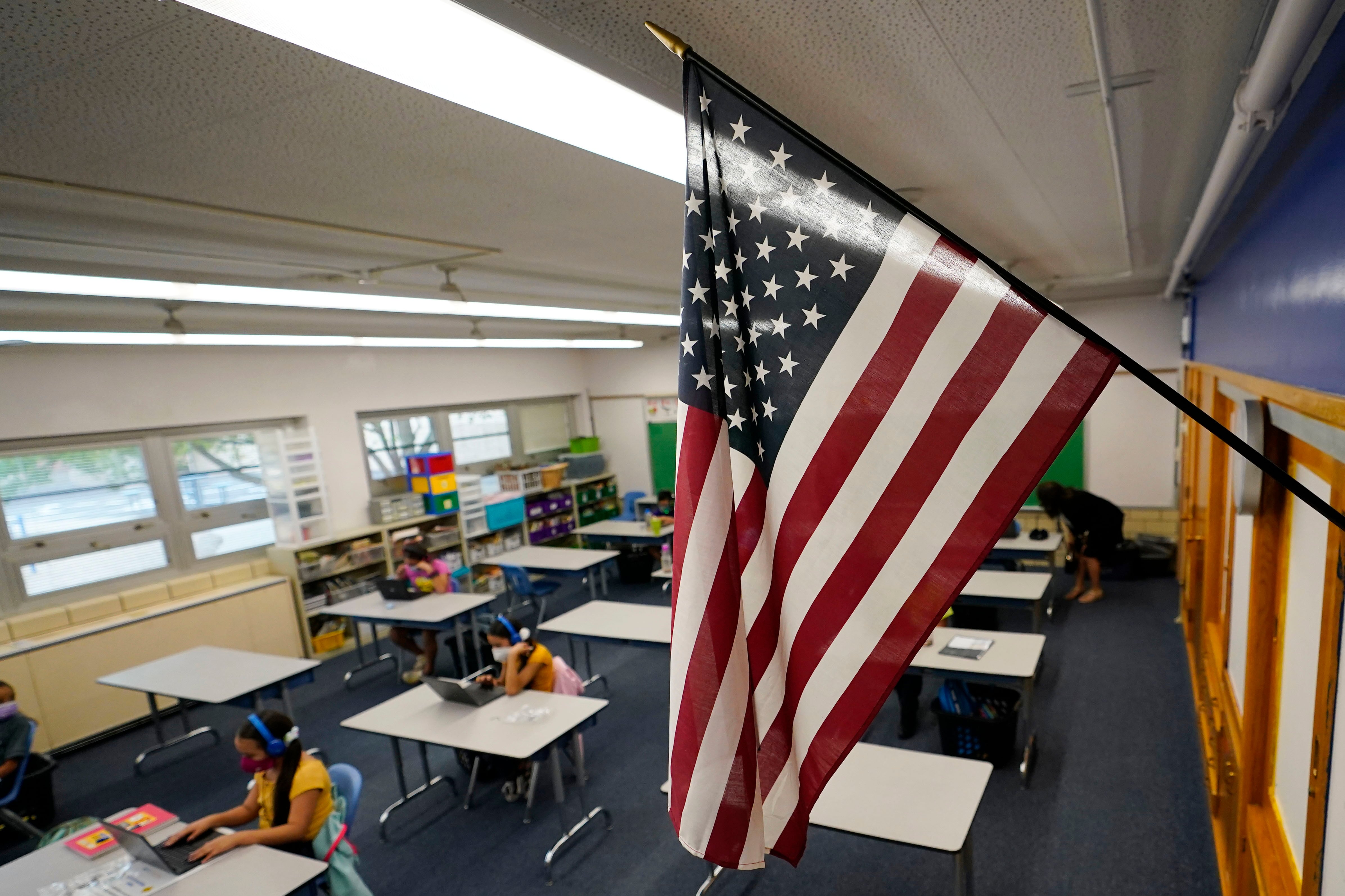 An American flag hangs over a classroom full of masked students working at desks.