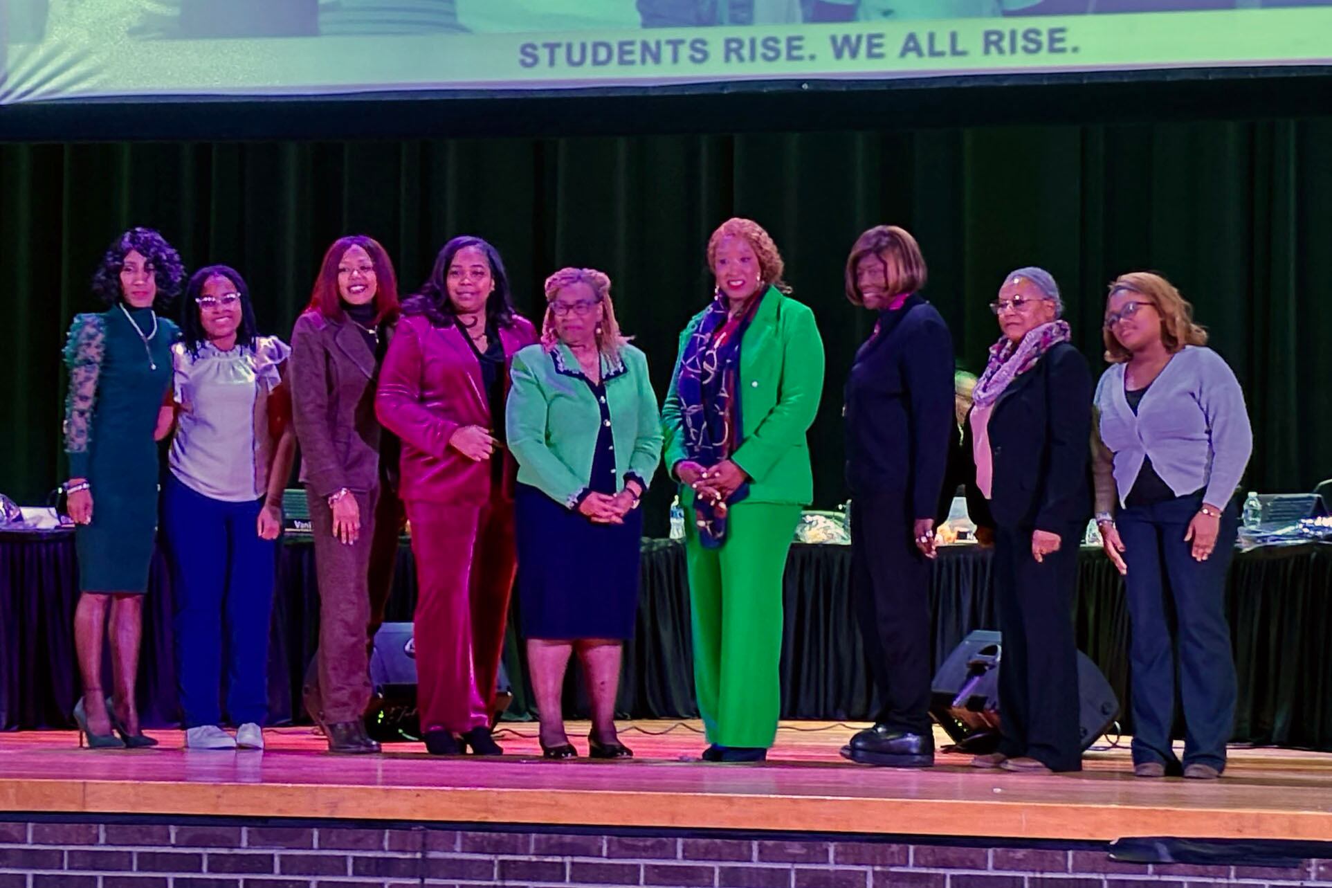 A group of women in business clothes stand in a line posing for a photograph on stage in front of a long table.