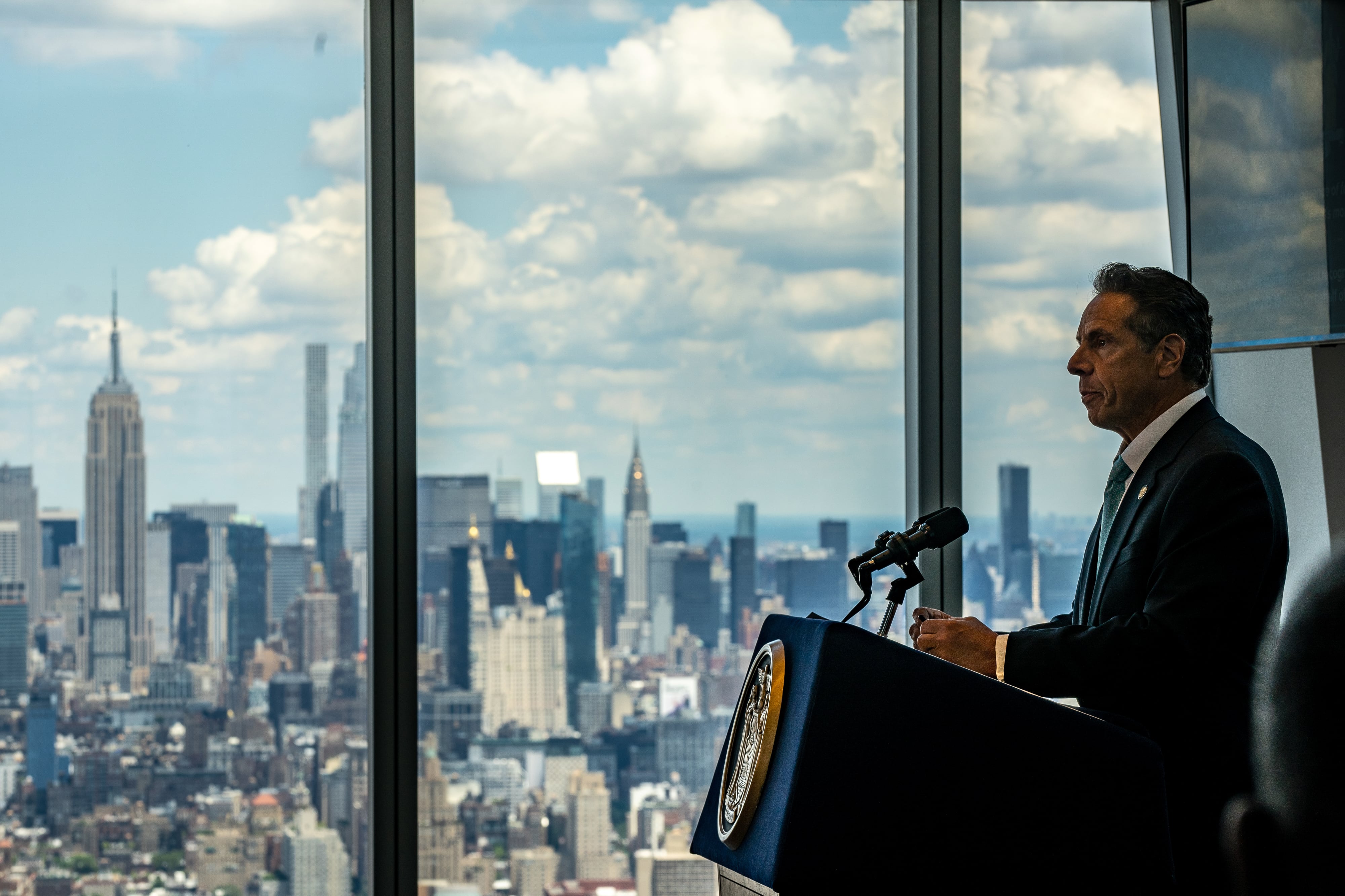 New York Governor Andrew Cuomo speaks at a podium indoors, with the New York City skyline visible behind the windows to his right.