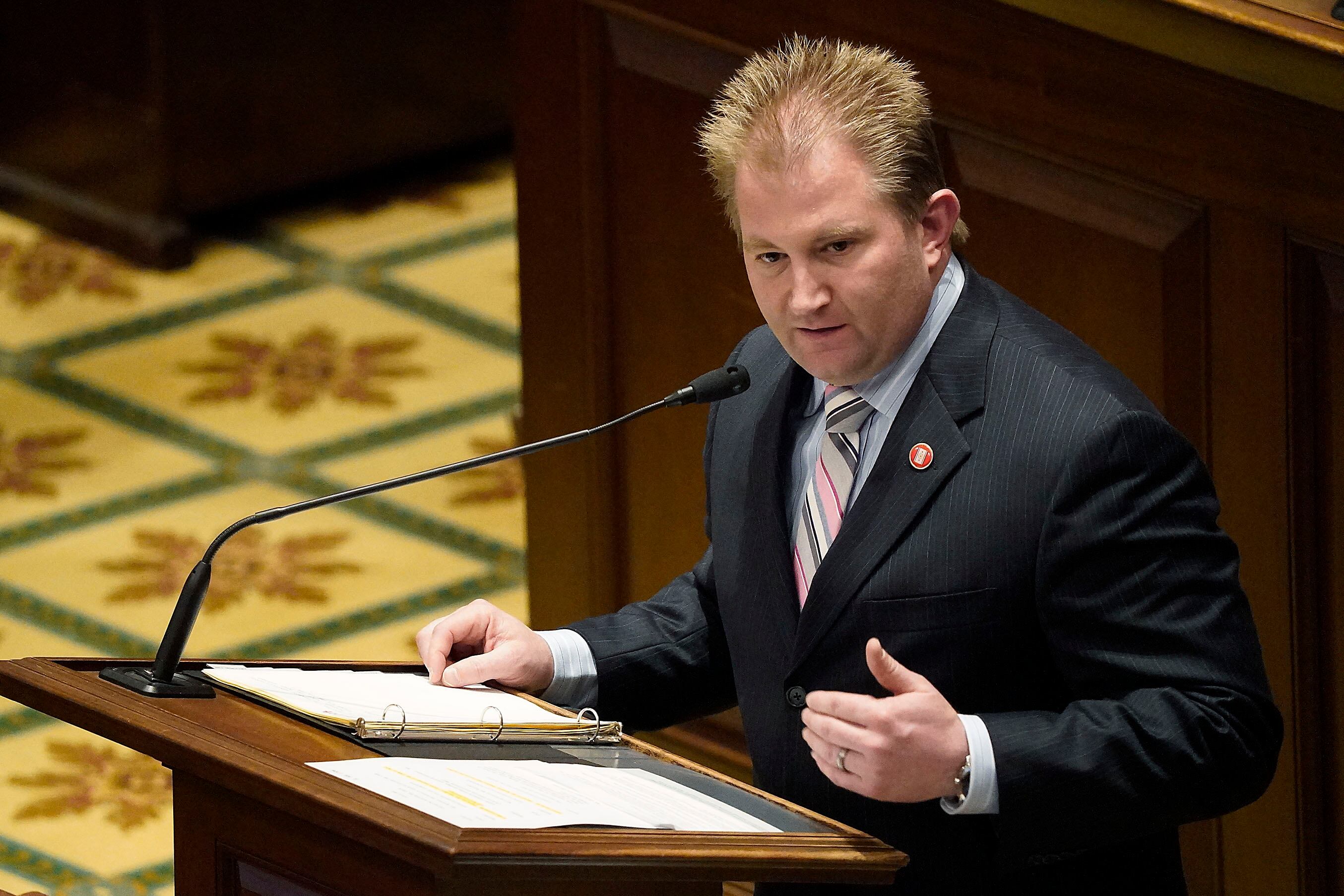 A man wearing a dark suit speaks from a wooden podium with yellow carpet and wooden panels in the background.