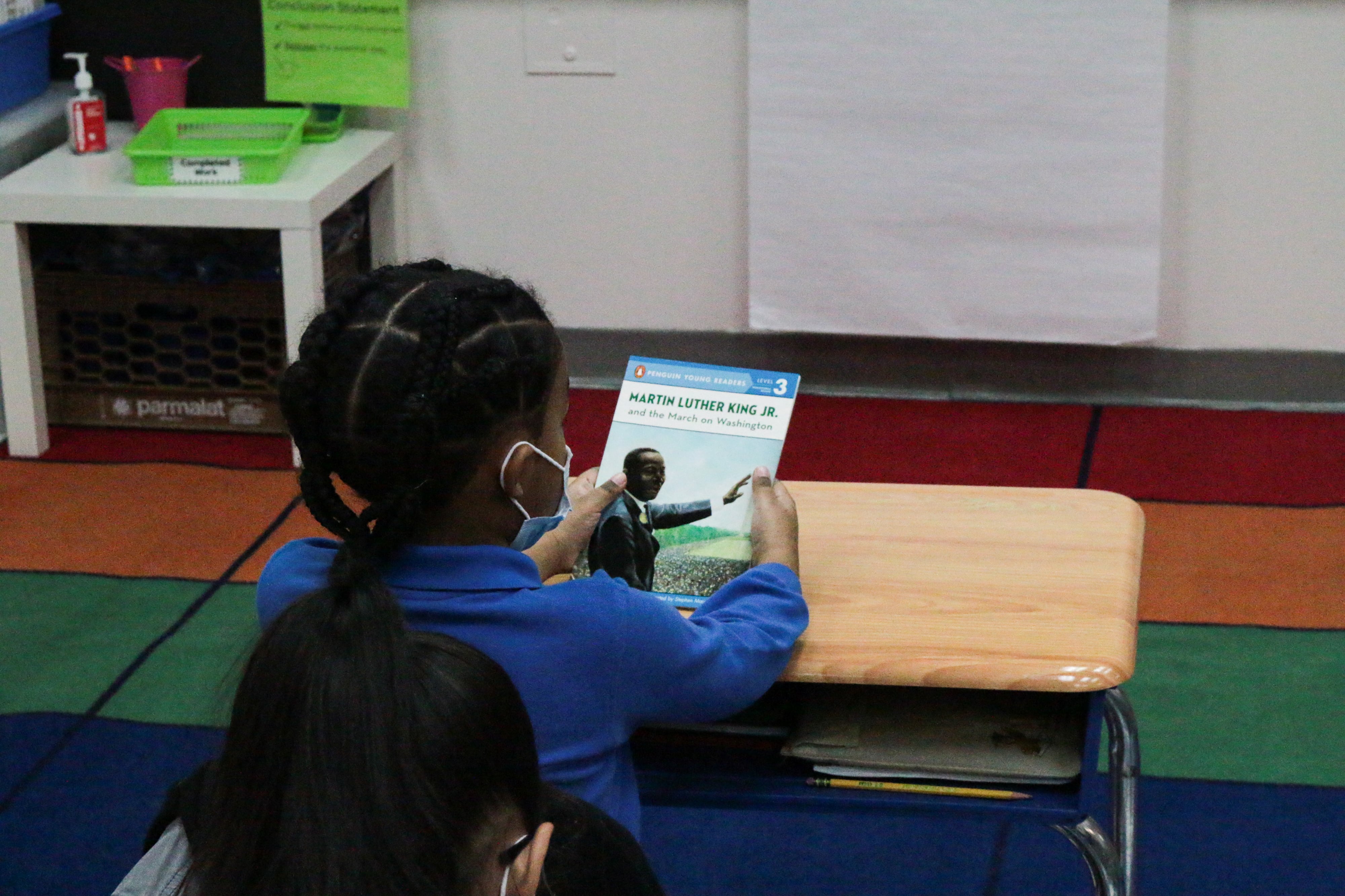 A student holds a book in front of them while they sit at their classroom desk.