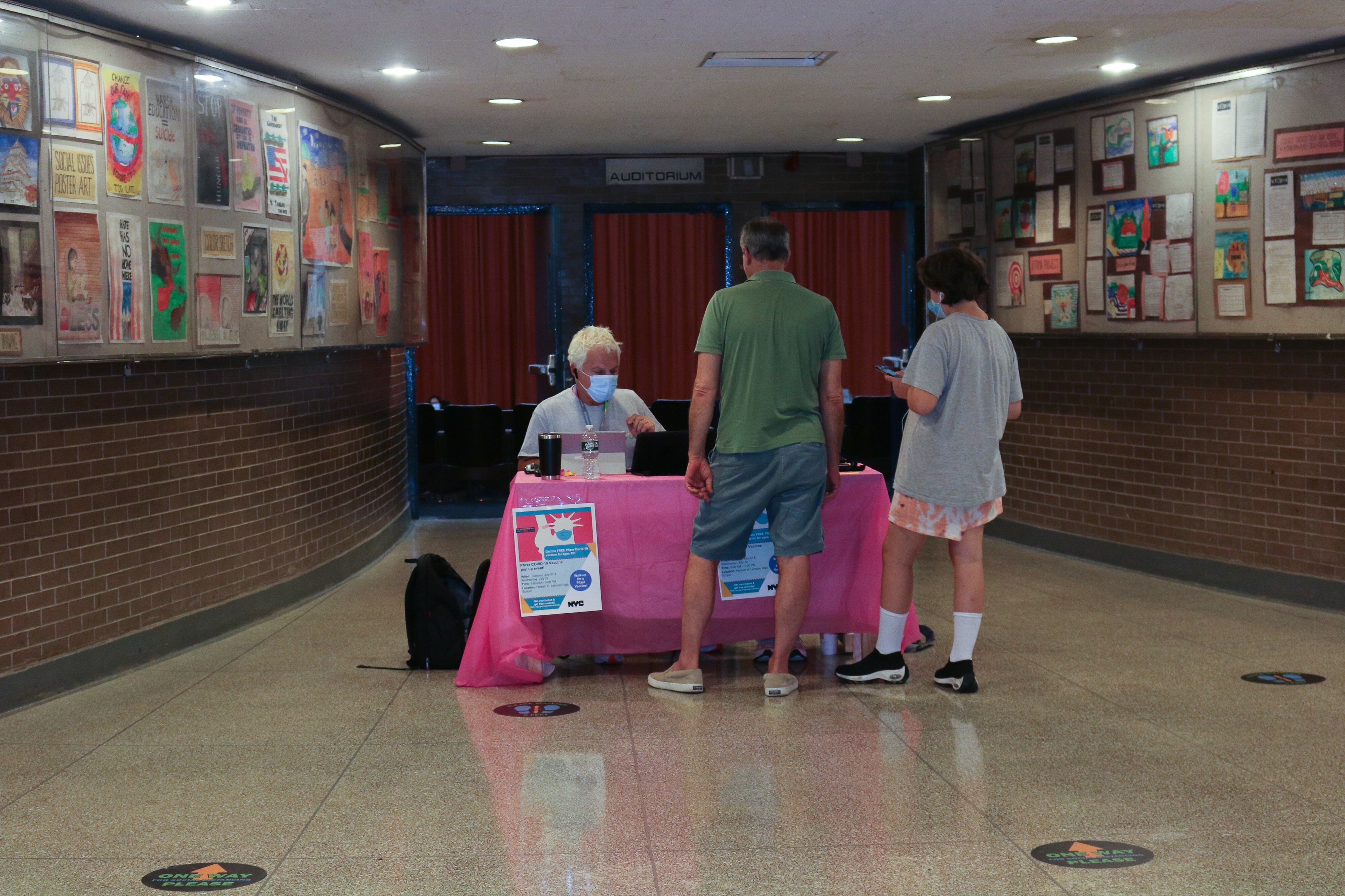 From a distance, two people stand in front of a table with a person sitting behind it.