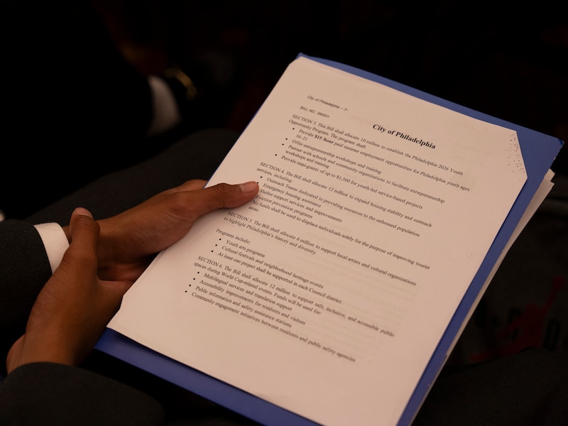 A photograph of a pair of hands holding a printed Philadelphia policy proposal with numbered sections and budget allocations.