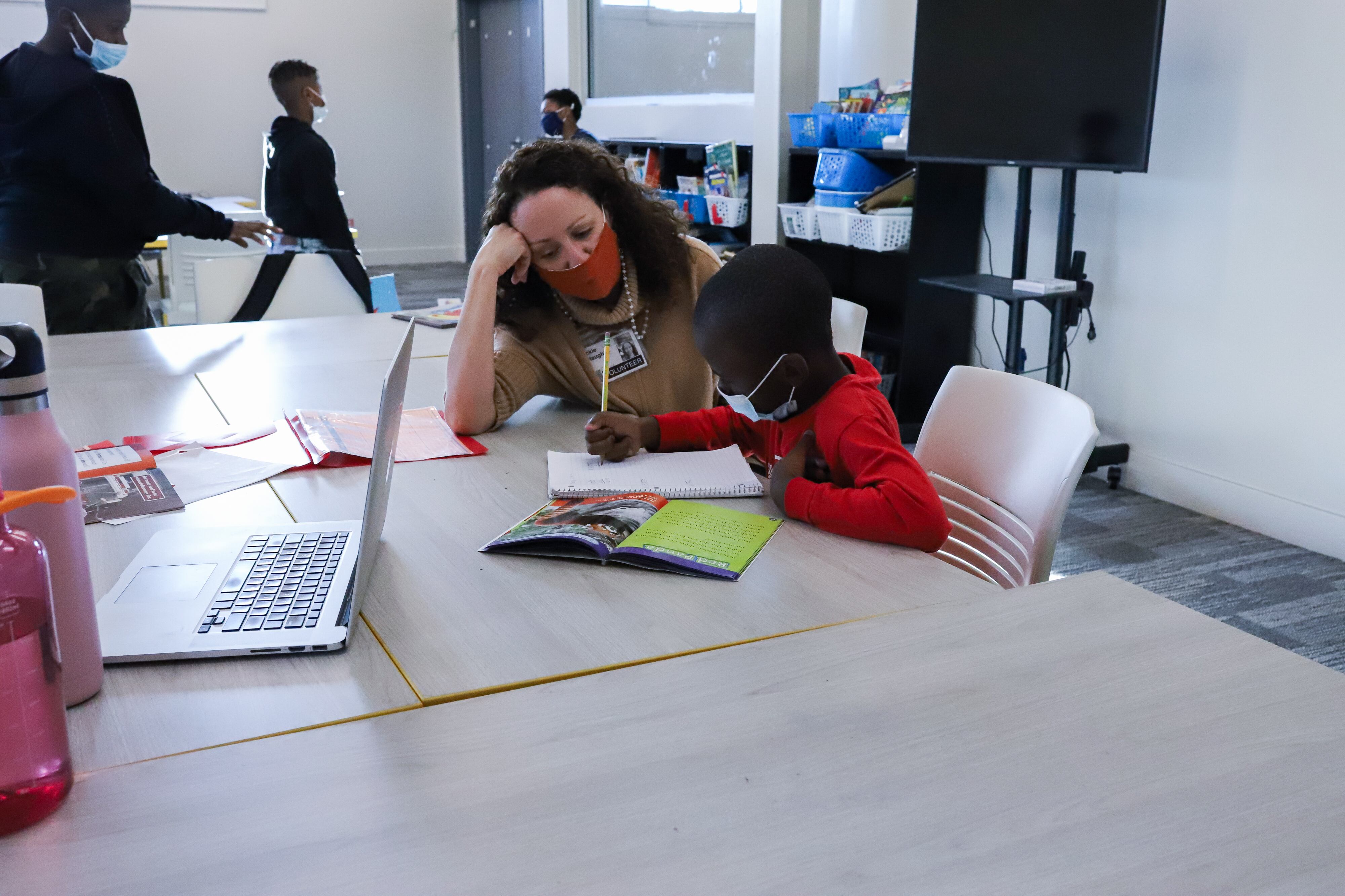 An educator works with a young student at a table as he writes in his notebook. Other students walk around the classroom behind them.