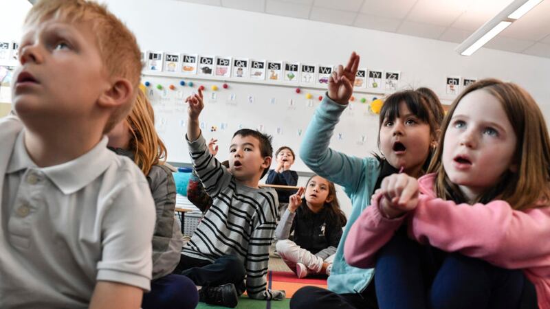 First-graders sit on a rug in a classroom. Their hands are in the air, tracing letters. Their mouths are open as they repeat after the teacher.