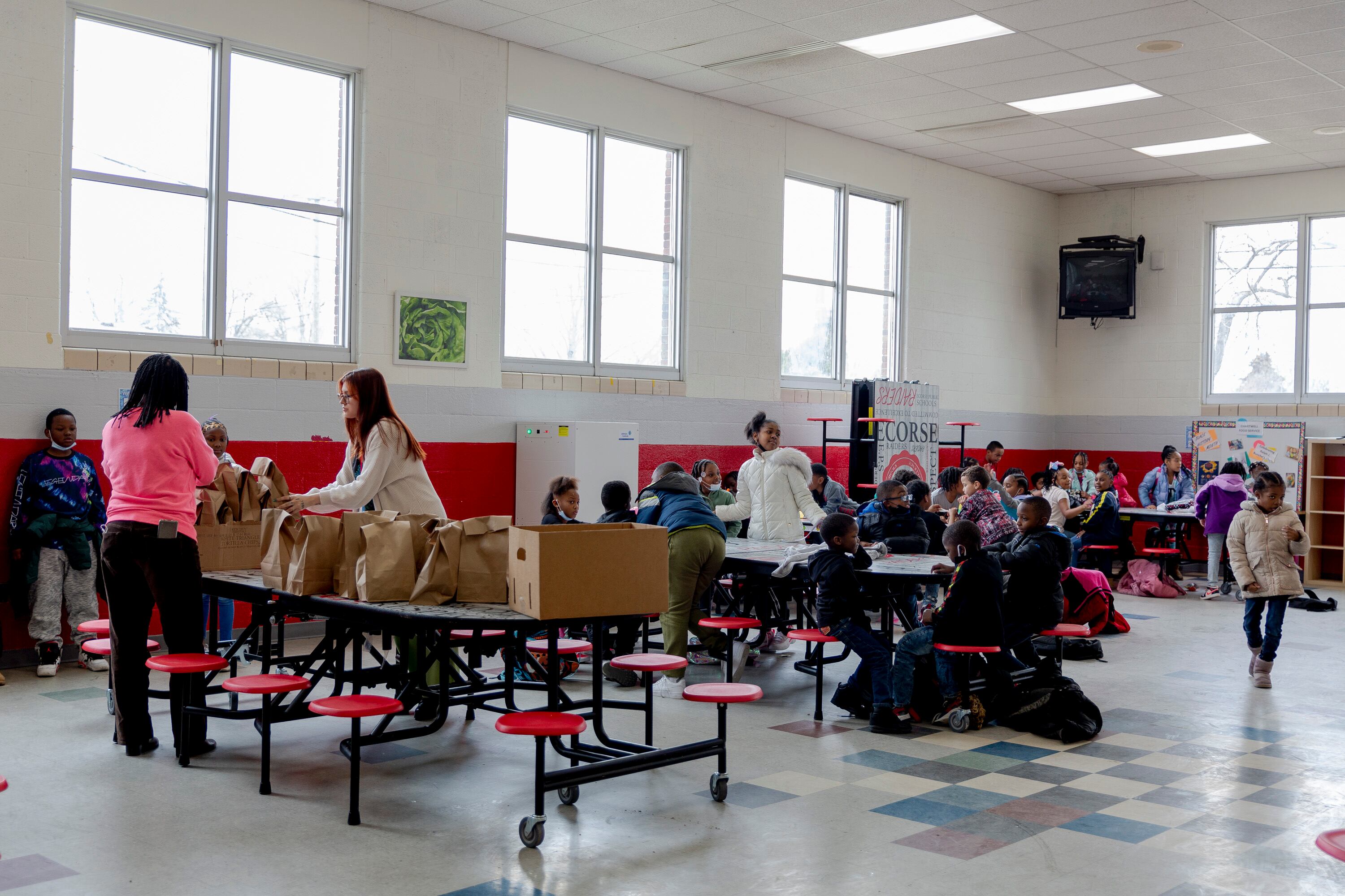 A large group of people sits at tables with red chairs in a cafeteria with linoleum tile floor and high square light-filled windows