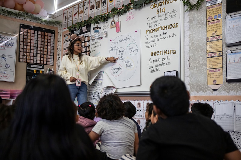 A woman in a white top and jeans points at a whiteboard in front of seated students.