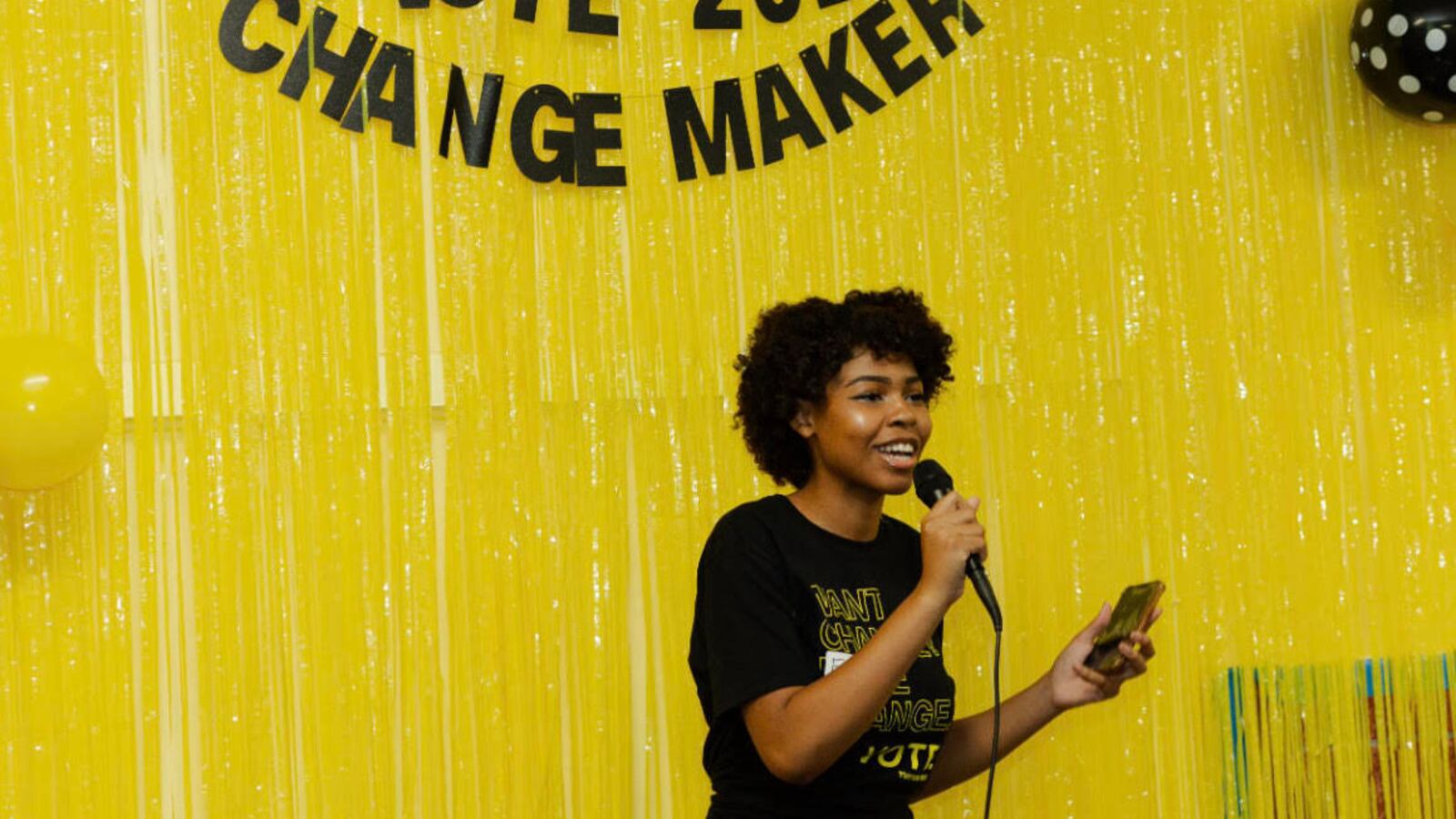 A young woman holds a microphone. Behind her, there is a yellow background that reads YVote 2023 Changemakers
