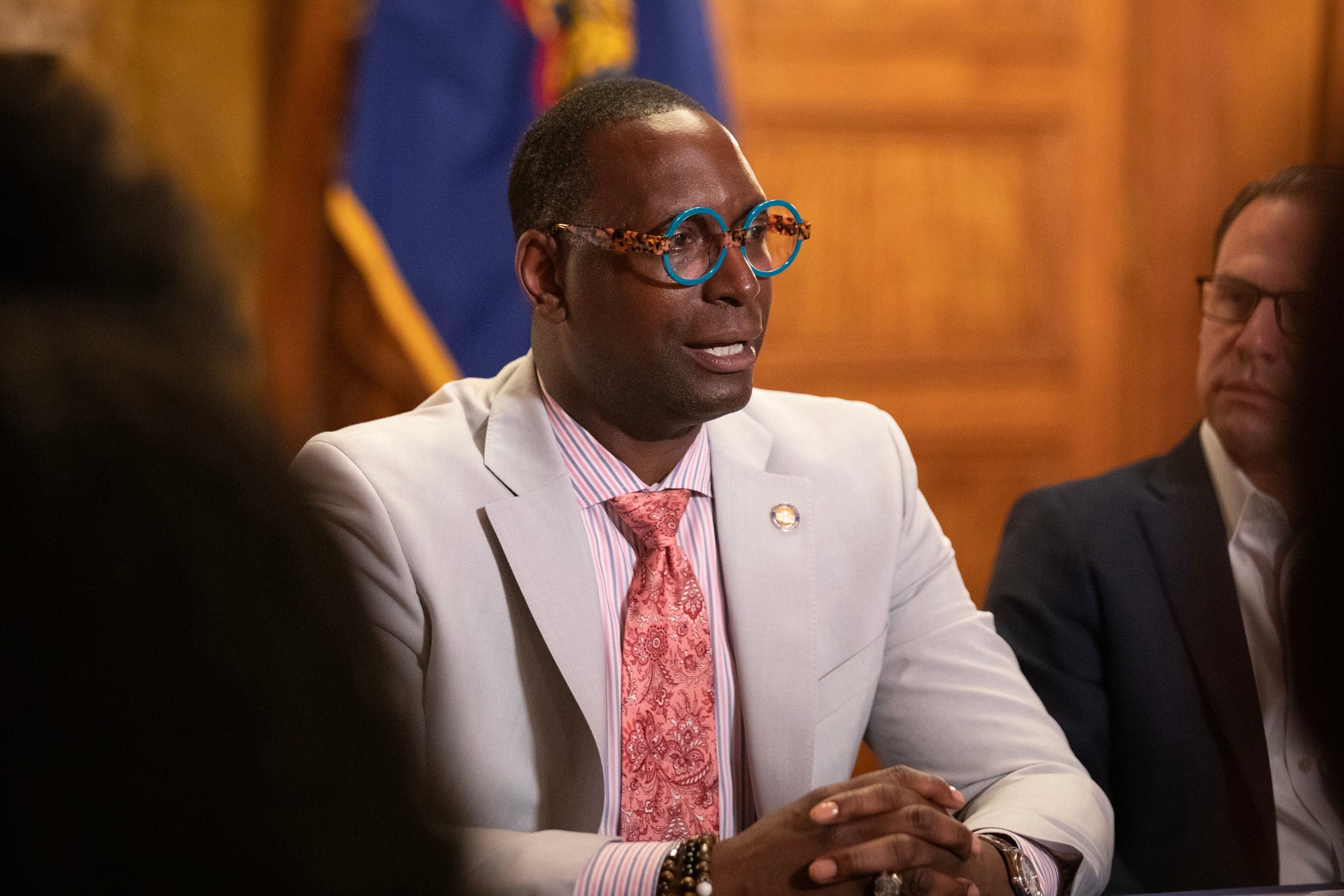 A man in a light suit jacket, blue glasses and a pink tie sits with his hands folded.