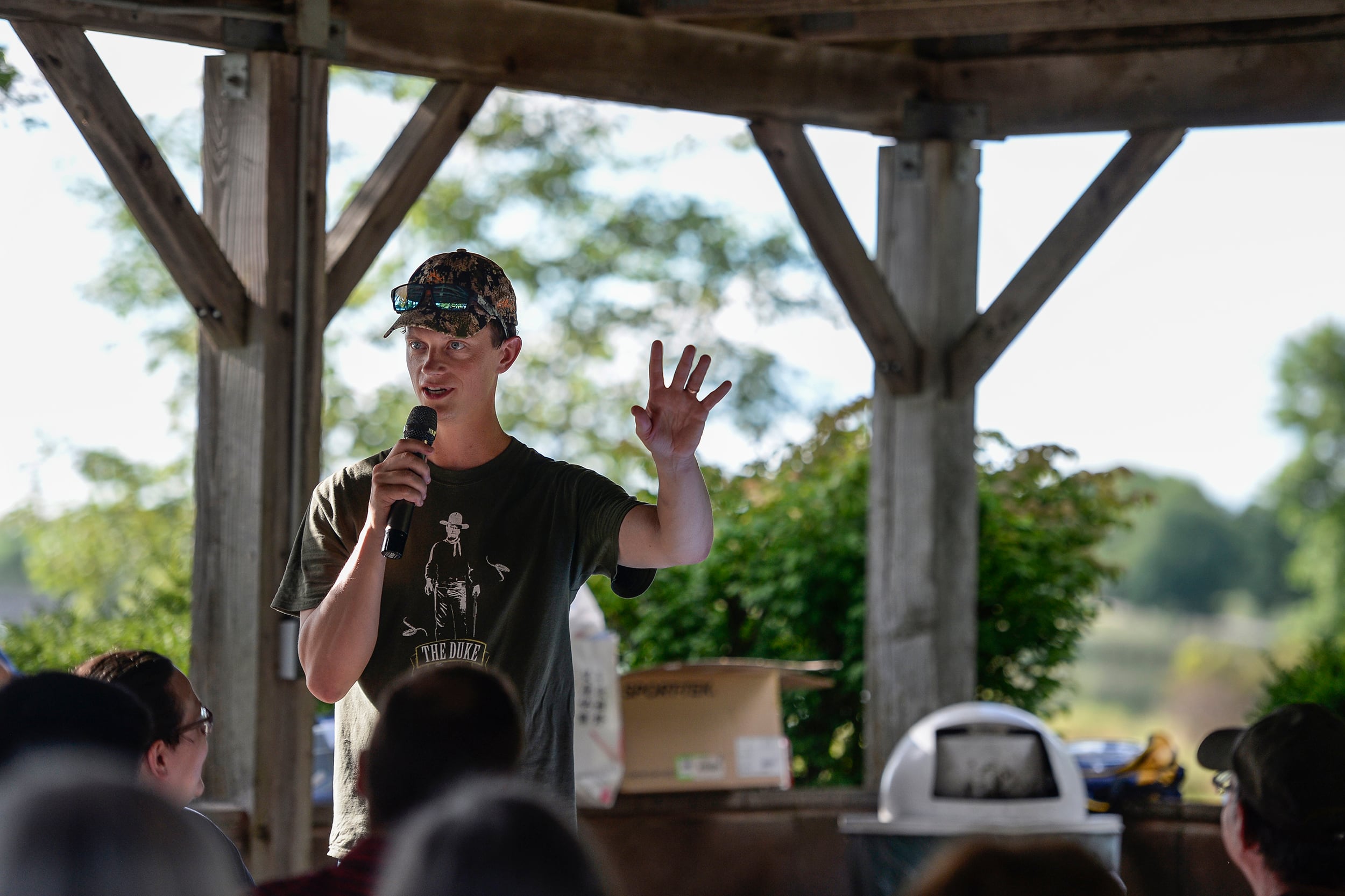 A photograph of a white man wearing a baseball cap and a t-shirt holding a microphone and speaking to a group of people underneath an outdoor wooden gazebo.