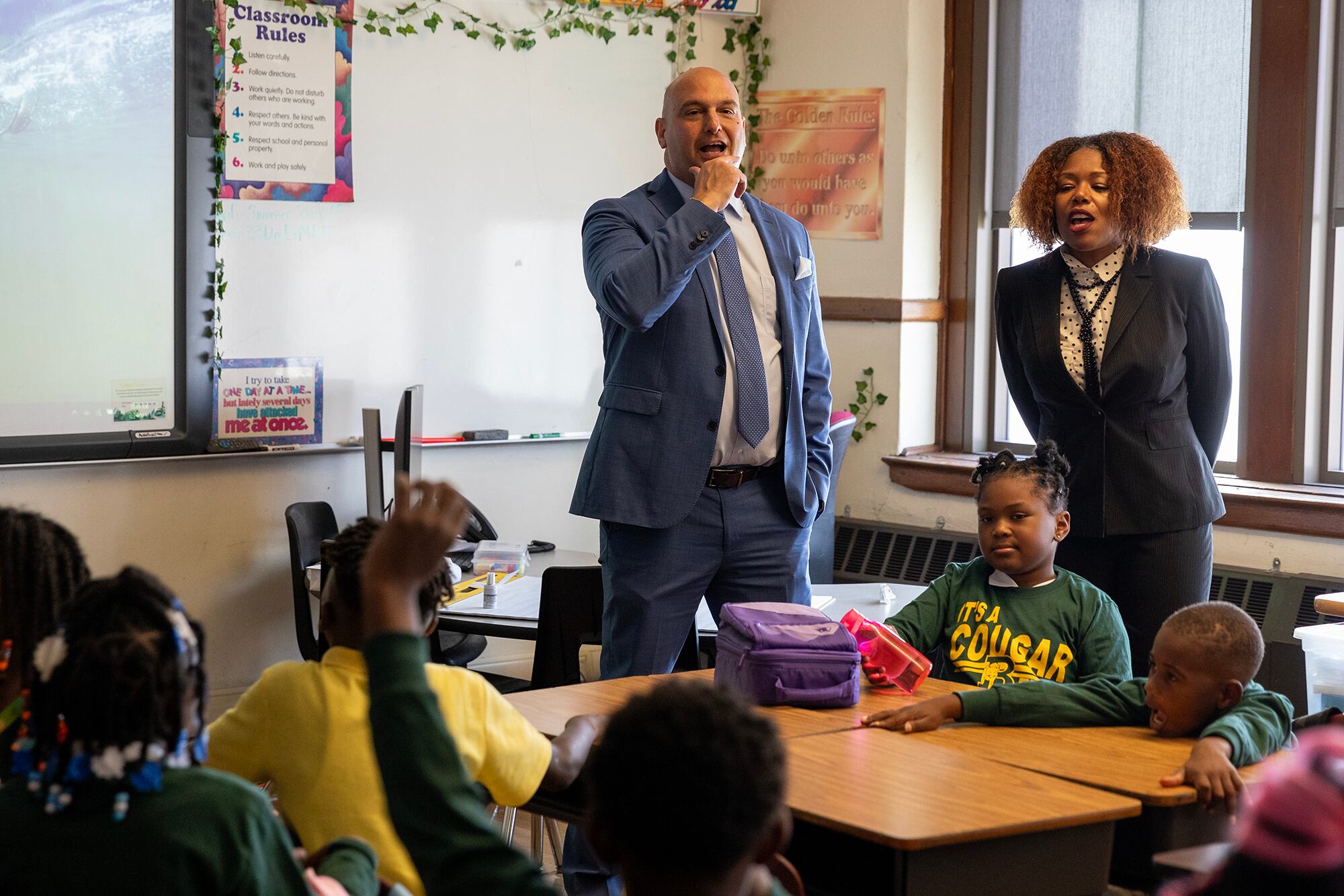 A man in a suit and a woman in a suit stand in a classroom, behind a group of students at a table.