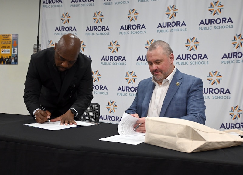 A photograph of two men in suits signing paper in front of a background.