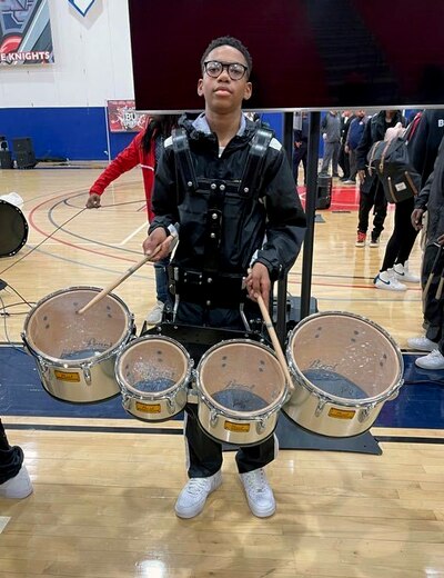A young boy wearing all black and a set of drums holds drumsticks while posing for a photograph.