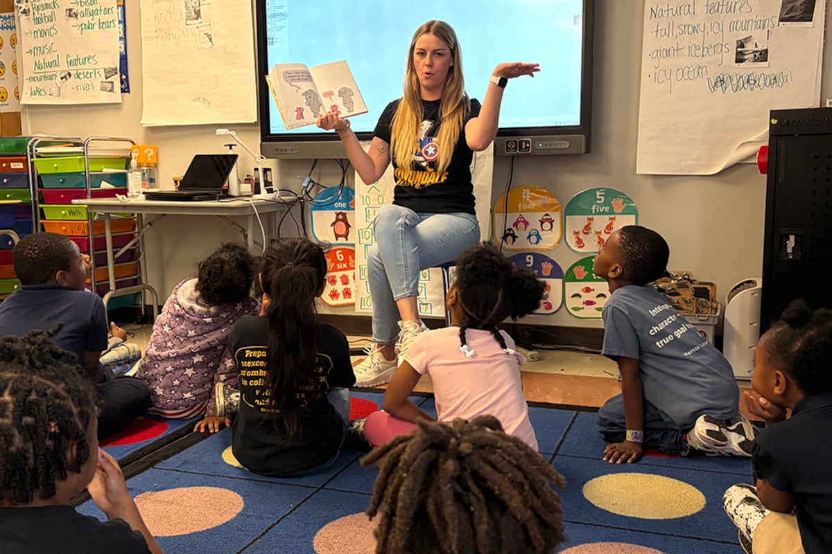 A woman with a black shirt gestures in front of a group of kids seated on a carpet.