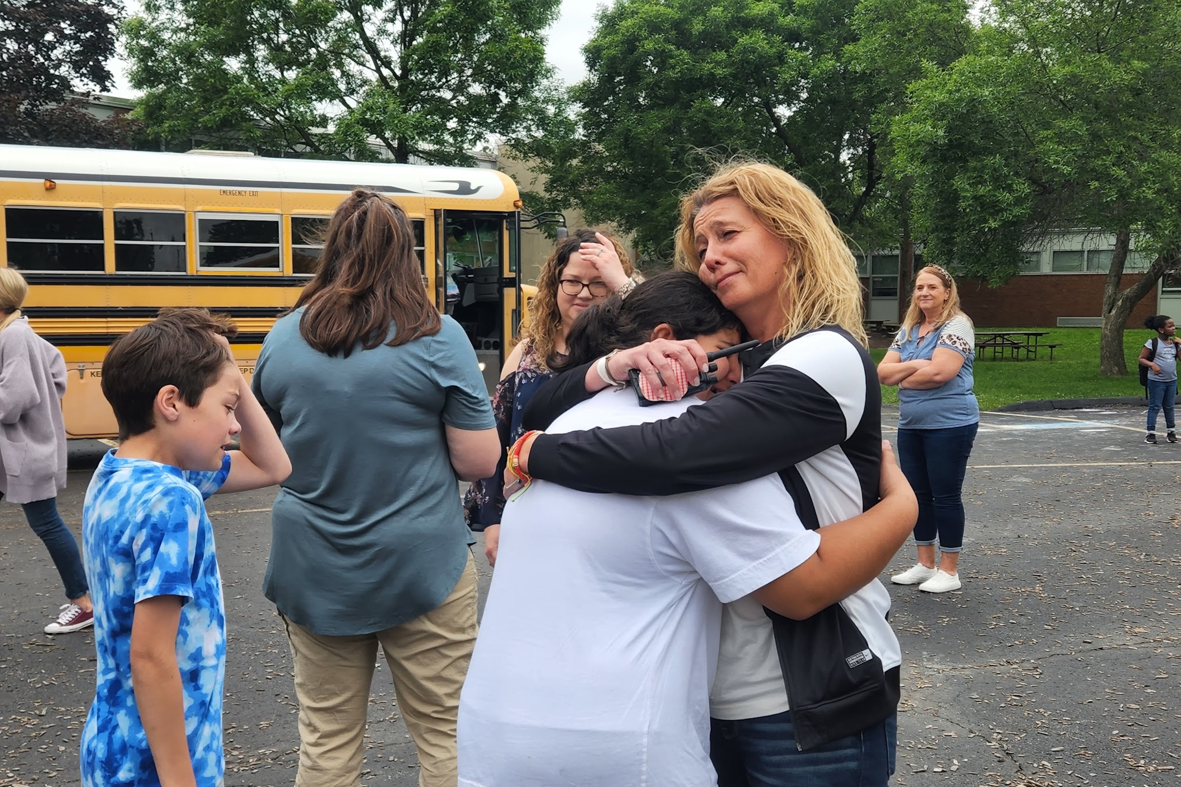A teacher hugs her student on a blacktop as other people walk behind them, in front of a yellow school bus.