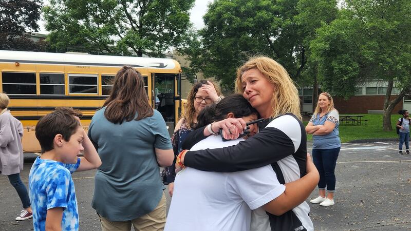 A teacher hugs her student on a blacktop as other people walk behind them, in front of a yellow school bus.