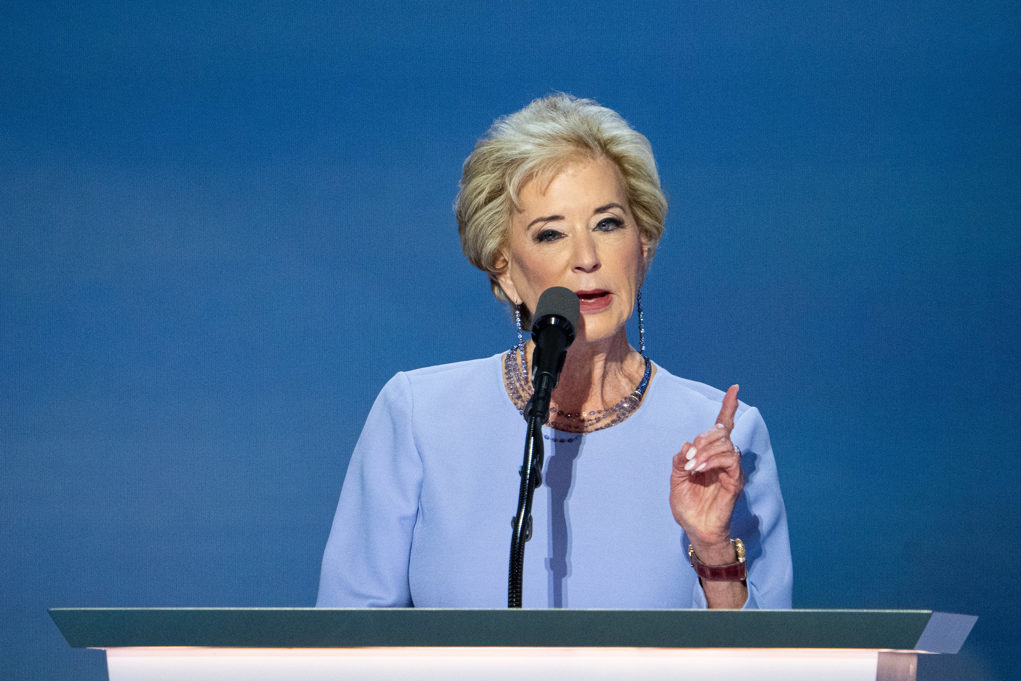A woman in a blue blouse speaks behind a black microphone and in front of a blue background.