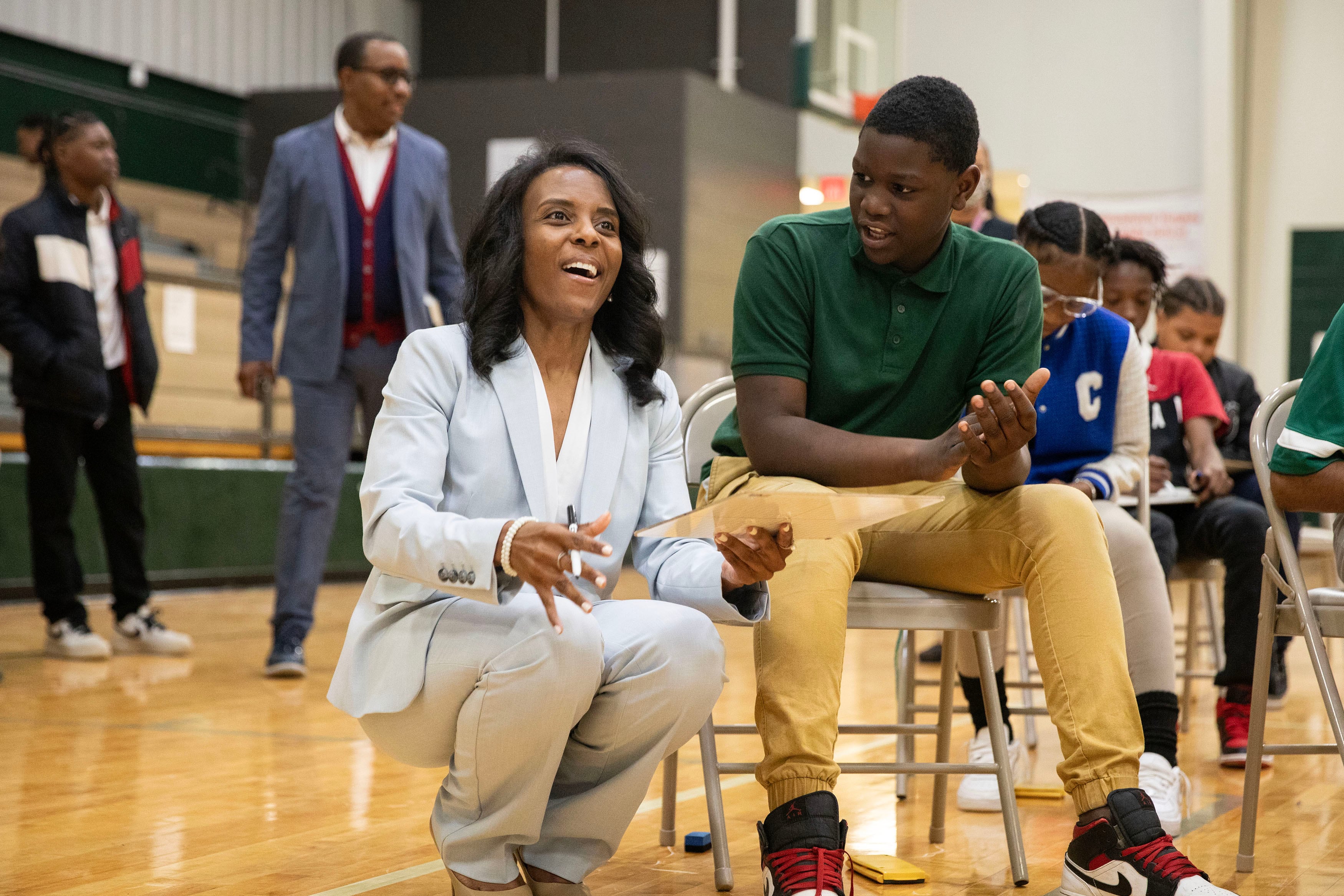 A woman with short dark hair and wearing a light blue suit kneels beside a young student wearing a green shirt and tan pants and sitting in a chair in a school gym.