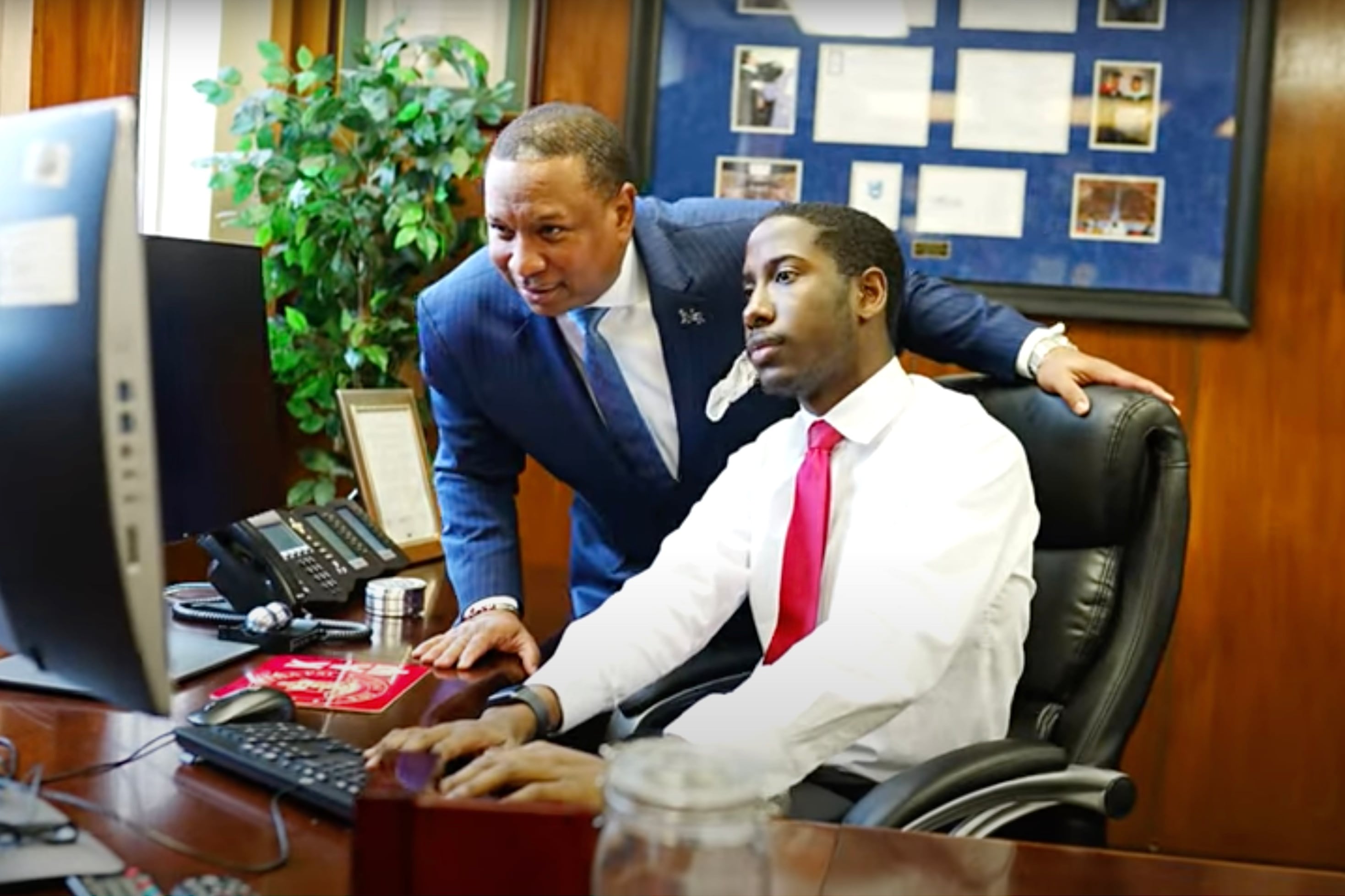 A man stands beside a male student sitting at a wooden desk, pointing at a computer