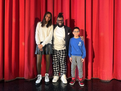 A teacher poses with two elementary students in front of a red curtain.