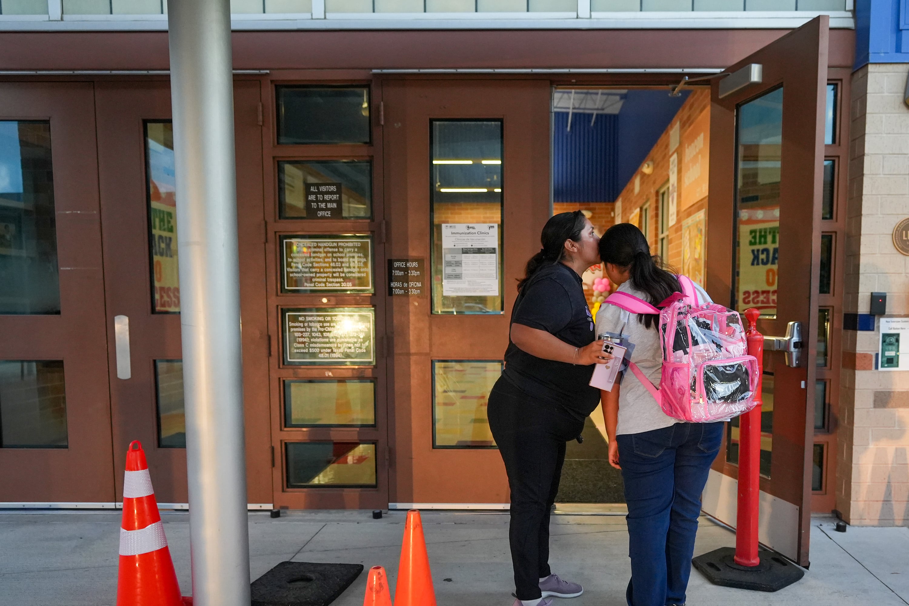 A woman in dark clothes kisses the head of a young girl with a backpack.