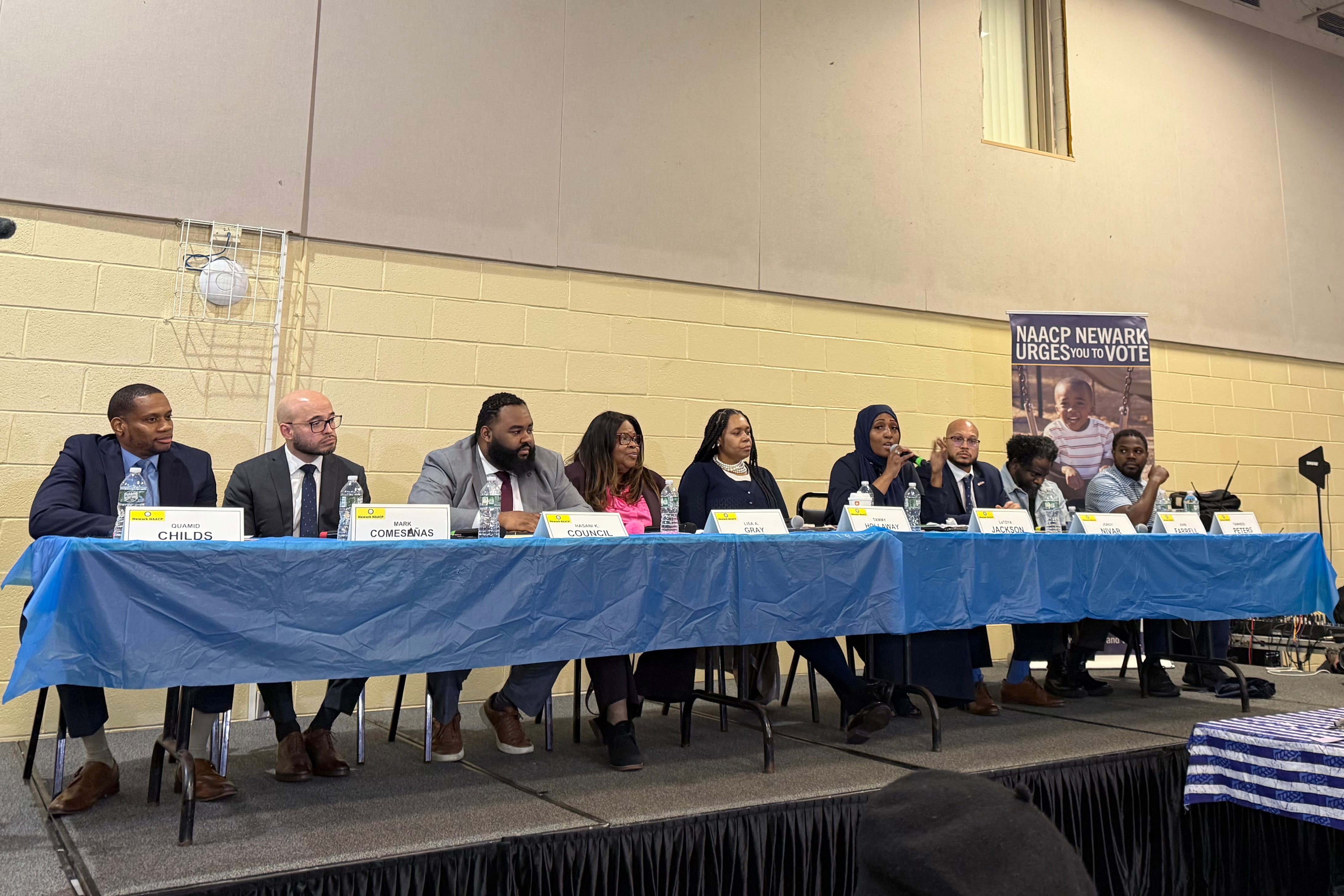 A photograph of a group of adults in business clothes sitting at a long table on a stage.